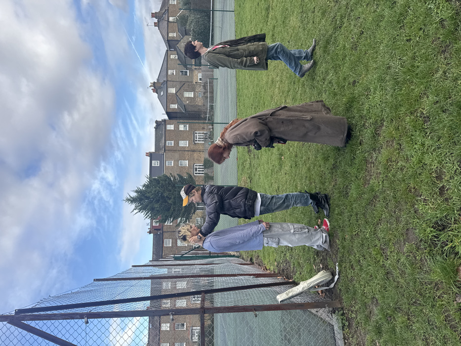 The image depicts an outdoor scene in a public park area adjacent to tennis courts, situated within a residential district of London, United Kingdom. Four individuals are present on a patch of green grass.
In the center-left, a male with blonde hair, wearing a light blue long-sleeved shirt and grey cargo trousers, stands facing right. Another male, dressed in a dark puffer jacket, blue jeans, black boots, and a yellow and white cap, stands directly in front of him. This second male has his right hand placed on the first male's neck/jawline and his left hand near the first male's hair, indicating an adjustment or styling action.
Immediately to their right, a female with reddish-brown hair, wearing a long brown trench coat and a patterned scarf, is bent forward at the waist, holding a black camera with a lens directed towards the two males, suggesting she is actively photographing or preparing to photograph them.
Further to the right, a fourth male with dark hair, wearing a green jacket over a light-colored shirt, blue jeans, and light grey shoes, walks from right to left, looking towards the left, not directly engaging with the other three individuals.
On the far left, leaning against a chain-link fence that borders the tennis courts, is a light-colored soft case or bag. The background features green tennis courts with white lines, behind which stand multiple-story brick residential buildings, typical of London architecture, with white-framed windows and dark roofs. A large evergreen tree is visible amongst the buildings. The sky is partly cloudy and blue, with a faint aircraft contrail visible in the upper right quadrant. The ground in the foreground is covered in green grass with some visible patches of bare earth.