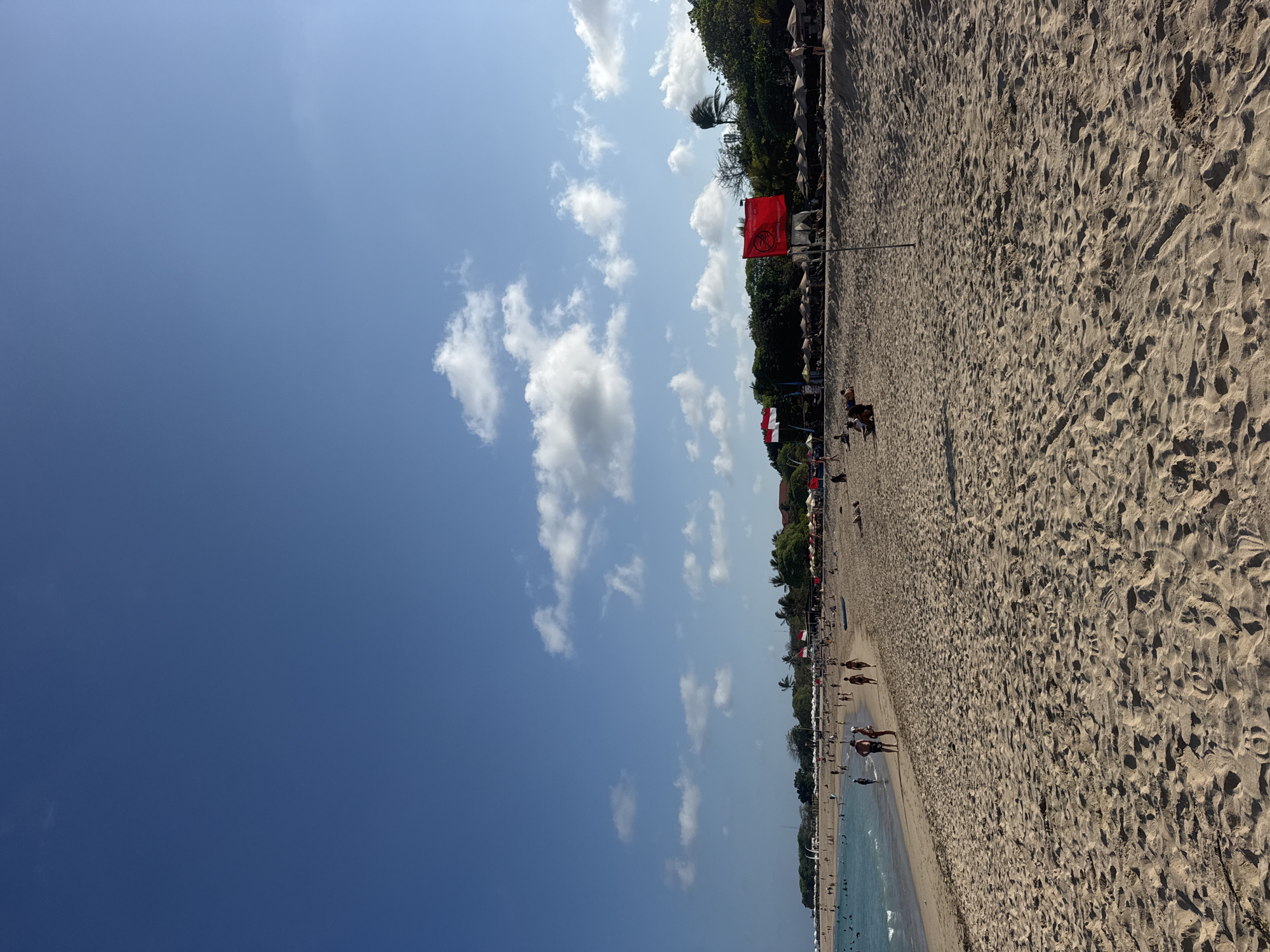 The image depicts a wide, sandy beach in Kuta Selatan, Indonesia, under a clear blue sky with scattered white clouds. The foreground is dominated by the textured sand with footprints. In the mid-ground, the turquoise ocean meets the shore, with gentle waves and several people visible in the shallow water and along the coastline. A diverse group of people are scattered across the beach, some walking, some sitting, and others sunbathing. Beach umbrellas and lounge chairs are set up in rows behind the beachgoers, suggesting a resort area. Two red and white flags, resembling the Indonesian flag, are prominently displayed, along with a red flag bearing a graphic symbol, possibly indicating a warning or prohibition, likely related to swimming conditions. Lush green trees and foliage form a backdrop along the upper edge of the beach, with some buildings visible amongst the trees. The overall scene conveys a lively and popular tourist destination.