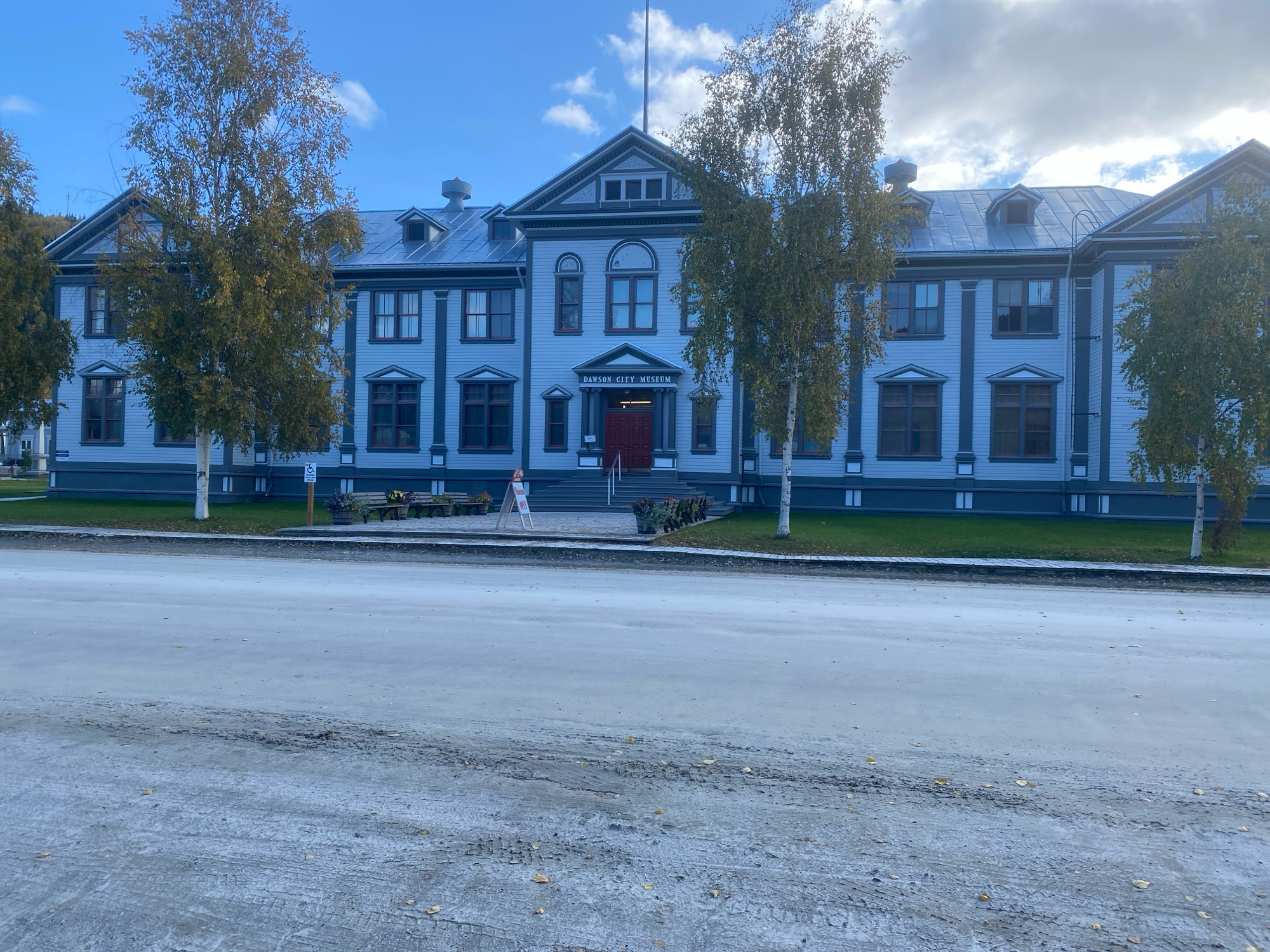 The image shows the exterior of the Dawson City Museum, located in Dawson City, Yukon, Canada. The museum is a two-story building with light blue siding, dark gray trim, and a dark gray roof. It has a symmetrical facade with several windows on each floor and a prominent central entrance with a red door. Above the entrance, a sign reads "DAWSON CITY MUSEUM".
In front of the museum, there are several birch trees with yellowing leaves, indicating it is autumn. A paved walkway runs in front of the building, leading to the entrance steps. To the left of the walkway, there is a sign with a wheelchair accessibility symbol. Benches and planters are visible in front of the museum.
The foreground of the image shows a dirt or gravel road with some tire tracks and scattered leaves. The sky is blue with some scattered clouds. The overall scene depicts a historic building in a northern town during the fall season.