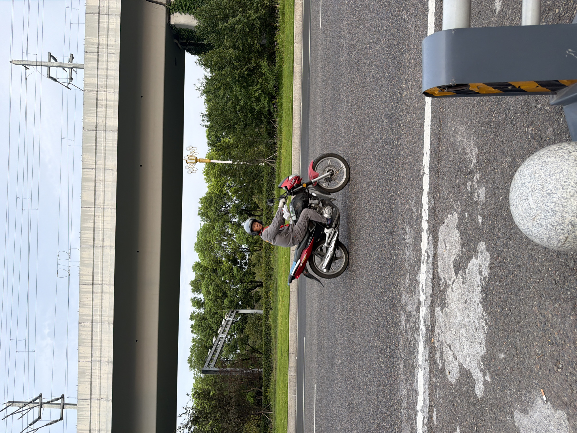 A man wearing a grey uniform and a helmet rides a red and black motorcycle on an asphalt road under a concrete overpass. The road has white lane markings. Green trees and a lamppost line the right side of the road. Overhead, power lines crisscross the sky. The scene appears to be in Jiangjin District, Chongqing, China.
