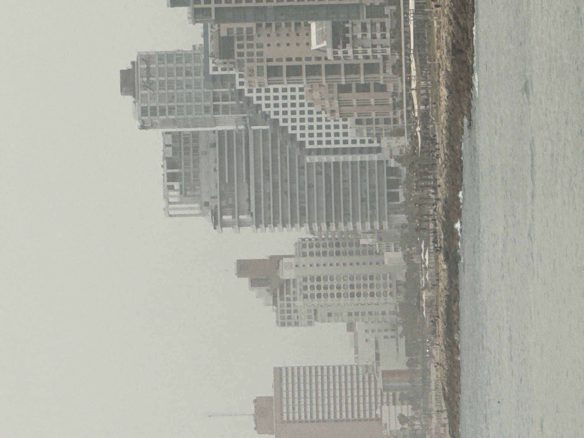 The image shows a hazy view of the Tel Aviv, Israel skyline along the Mediterranean coast. Several tall buildings are visible, appearing as concrete and glass structures with grid-like windows, suggesting a modern architectural style. The sky is overcast, which mutes the colors and reduces visibility. The shoreline is visible, with a beach and a concrete barrier. The sea is calm and reflects the hazy sky. The scene implies a sense of urban coastal development in a city with high population density.