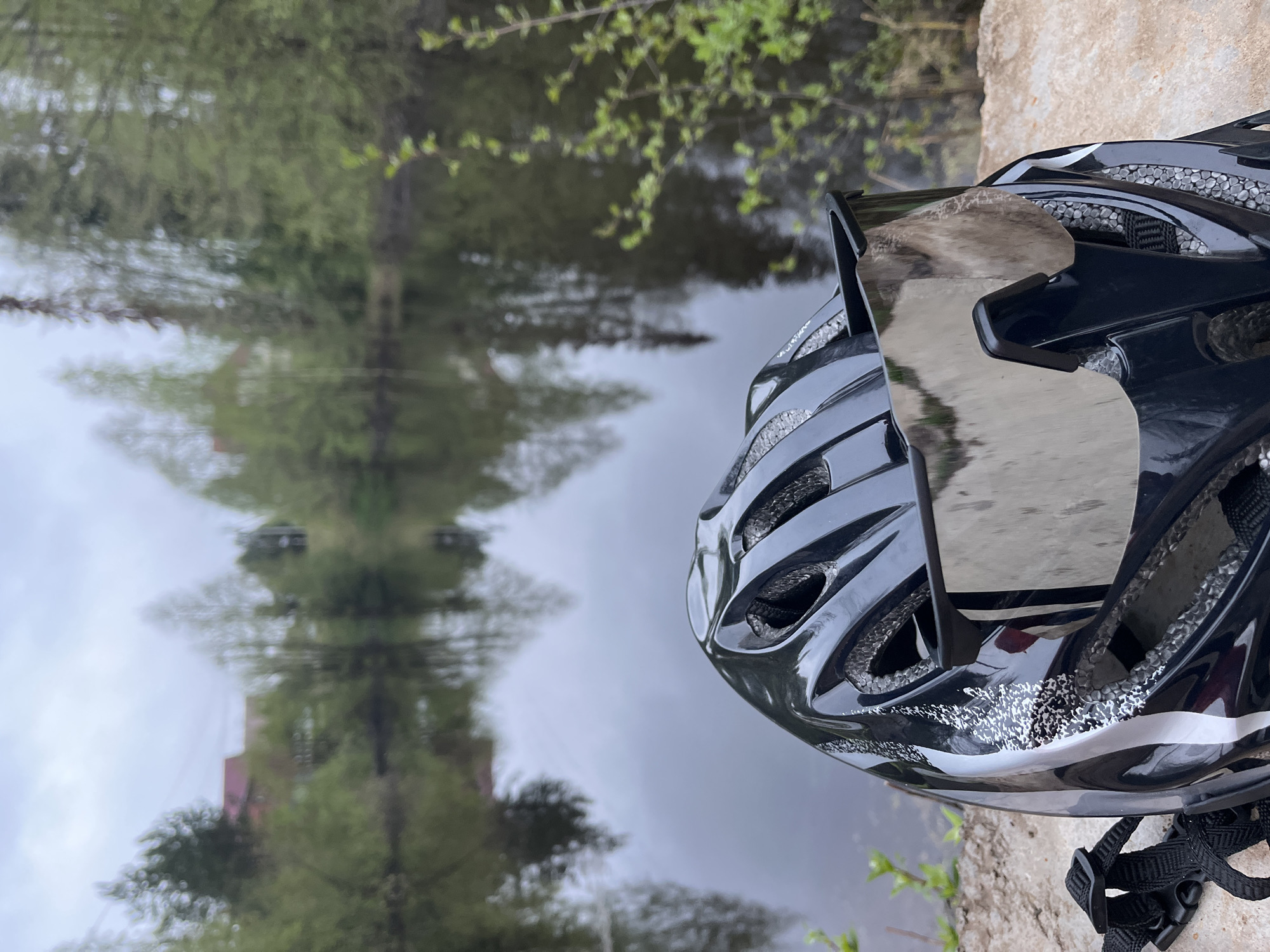 A close-up, angled shot features a black and silver cycling helmet in the foreground, positioned against a backdrop of a body of water reflecting trees and what appears to be a building. The helmet is detailed with ventilation holes and adjustable straps. Behind the helmet, the water reflects a blurry, upside-down image of green trees and a muted red structure. The location is the city of Chaikovsky, Russia, as inferred from the user’s prompt.