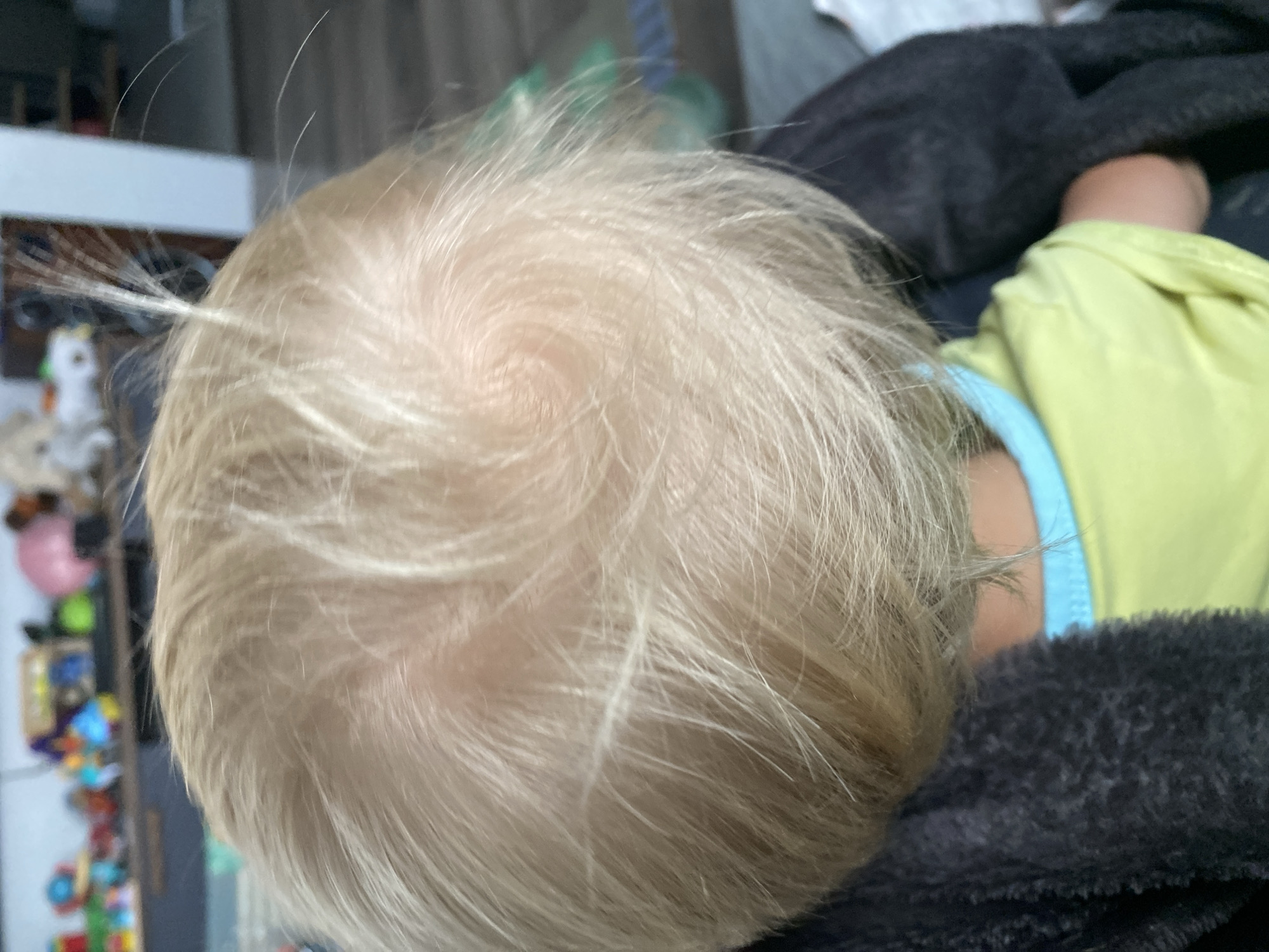 The image, taken from a top-down perspective, shows the back of a young child's head, focusing on their blonde hair. The hair is fine and appears to have a swirl pattern at the crown. Some individual strands stand out, particularly a few at the top that are sticking up. The child is wearing a light green t-shirt with a light blue trim around the neckline. A dark, fuzzy blanket is visible draped over the child's shoulders and back. In the blurred background, a shelf with various colorful toys is discernible, as is part of a white wall. The scene appears to be indoors, possibly in a home environment.