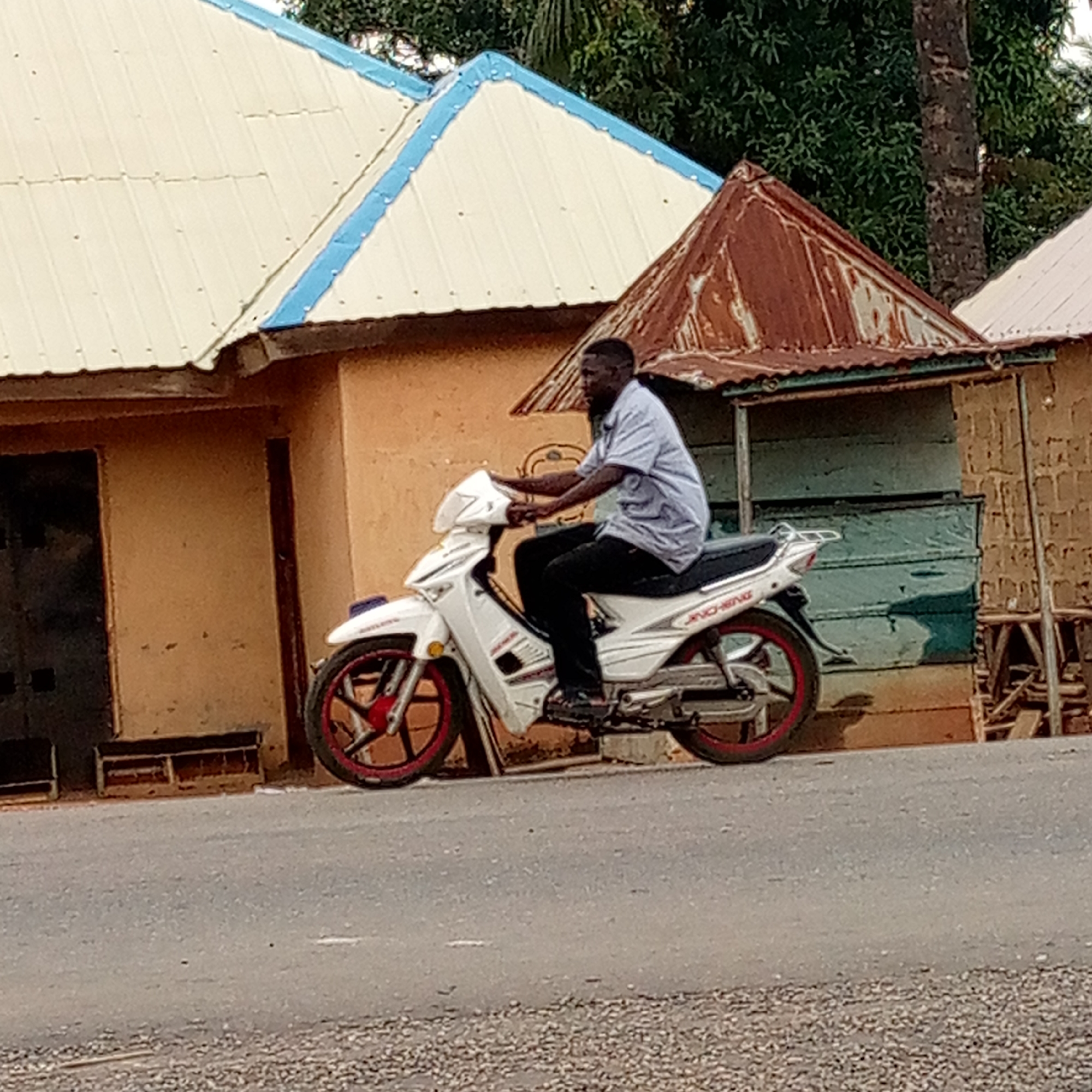 The image shows a man riding a white motorcycle with red rims on a paved road in Mararaba, Nigeria. The motorcycle is parked next to a building with an orange facade and a roof with a light-colored top and blue trim. To the right of the motorcycle is a small, shed-like structure with a rusty roof and green walls. The road surface is cracked and has a gravel shoulder. A person is riding a motorcycle. Buildings and green trees are in the background.