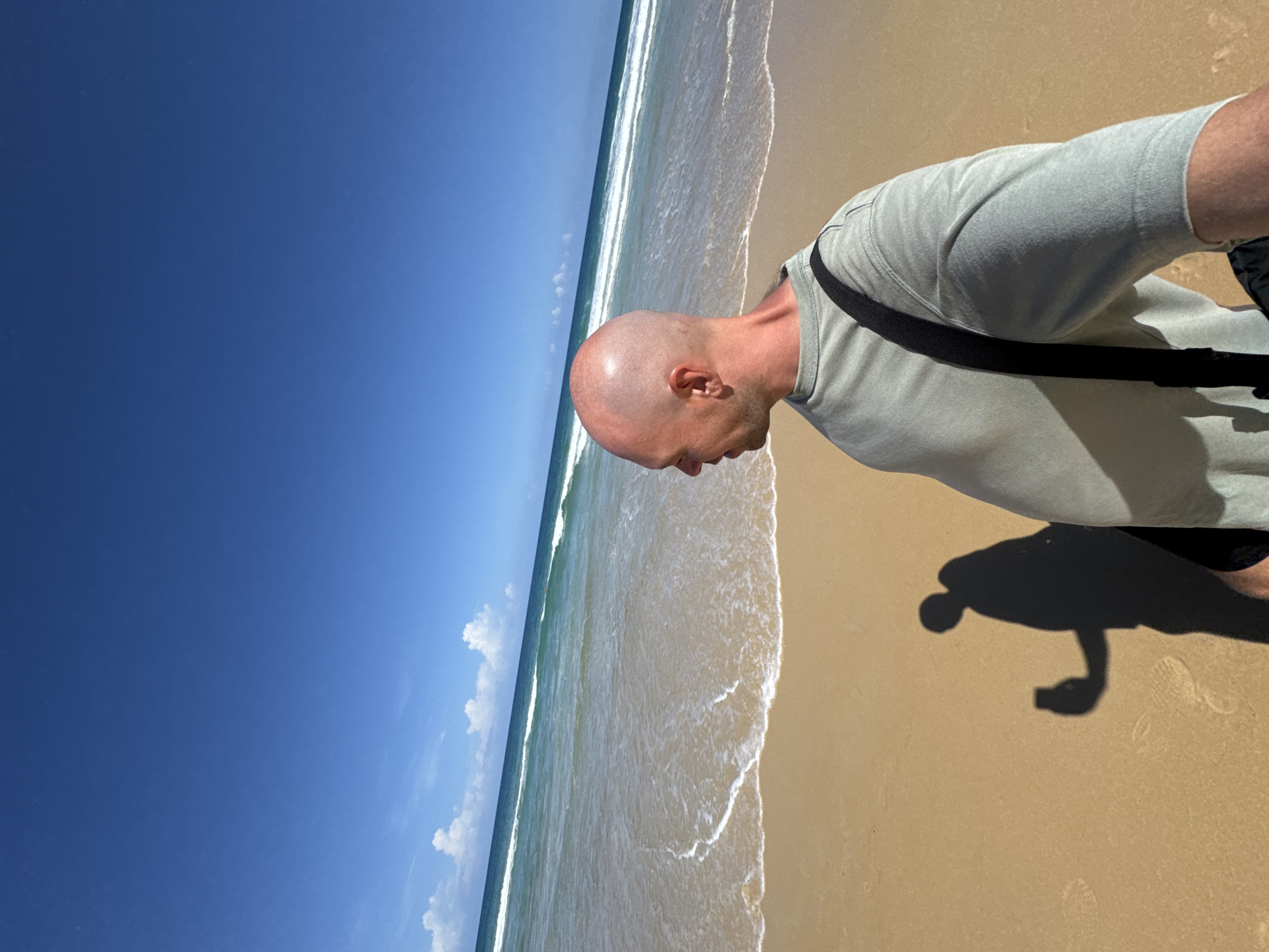 A bald man, wearing a light green t-shirt and a black bag strap across his shoulder, stands on a sandy beach. He is looking to the left, with the ocean stretching out before him. The water is turquoise with gentle waves and white foam. The sky is a clear, bright blue with a few wispy clouds. The man's shadow is cast on the sand to his left, and he is holding what appears to be a smartphone in his hand. The scene is bathed in bright sunlight, indicating a warm, tropical day. The location is Karon Beach, Karon, Thailand.