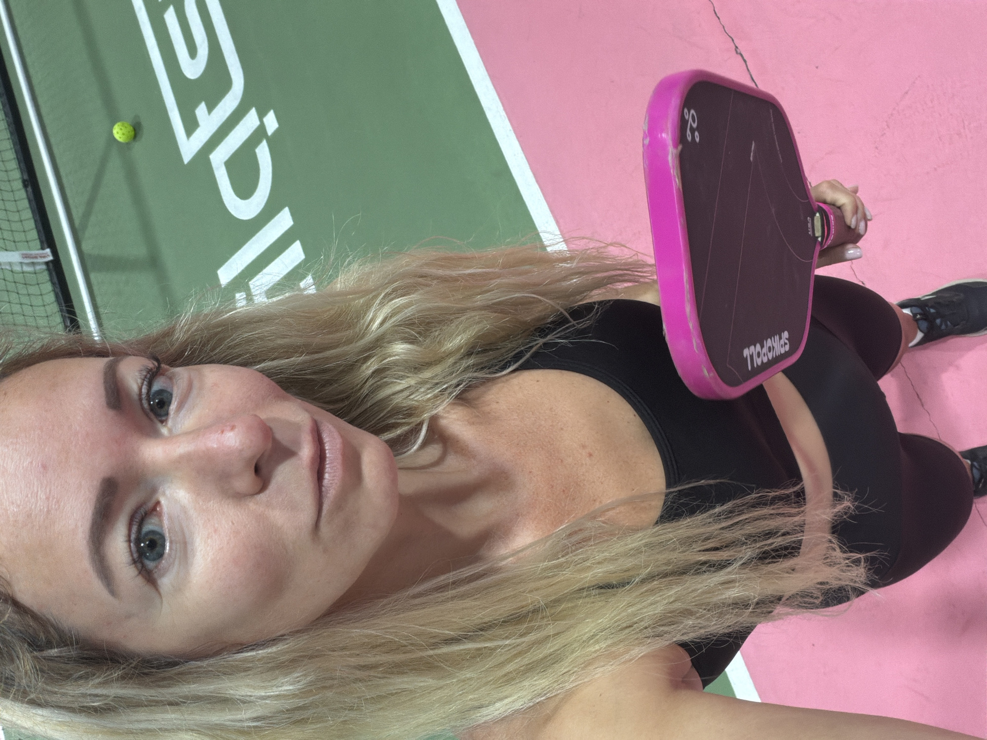 A woman with blonde hair and blue eyes is taking a selfie on a pickleball court. She is wearing a black sports bra and black shorts. She is holding a pink pickleball paddle in her right hand. The court is green and pink, with a white line dividing the two colors. There is a pickleball on the court in the background. The woman is looking at the camera with a neutral expression. The location is not Da Lat, Vietnam.