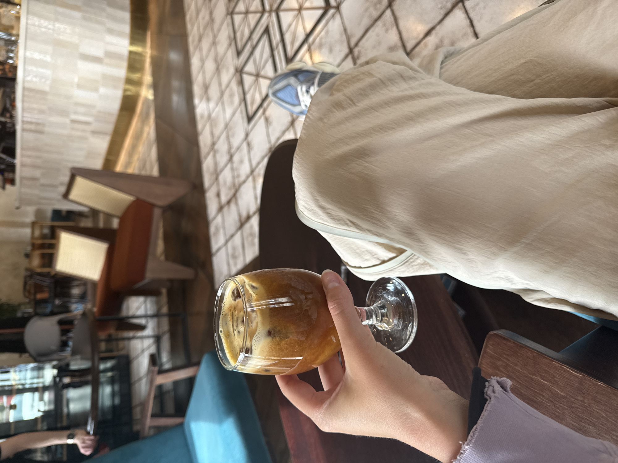 The image captures a first-person perspective of someone seated in a modern cafe in Krasnoyarsk, Russia. The main subject is a clear, stemmed glass held by a hand, containing a refreshing amber-brown coffee drink, likely an espresso tonic or cold brew, topped with a frothy head, ice, and small dark specks, possibly coffee beans or grounds. The hand holding the glass has neat nails.
The person is wearing light-colored, possibly beige or off-white, wide-leg trousers or a skirt, which show soft folds. Their right foot, clad in a light blue and white sneaker, is visible against a geometrically patterned tiled floor. A light purple sleeve with a rough-cut cuff, belonging to the person, rests near a dark wooden table edge in the foreground.
The background reveals a stylish cafe interior. The floor is covered in light-colored, geometric tiles with darker grout lines. Modern cafe furniture, including chairs with brown wooden frames and some with blue upholstered seats, is visible. A prominent design feature is a curved, reflective, brass or gold-toned wall or partition that separates different areas, with a light-colored tiled wall behind it. The lighting suggests it is daytime. The scene conveys a moment of relaxation or enjoyment in a contemporary urban setting. There is no visible text in the image.