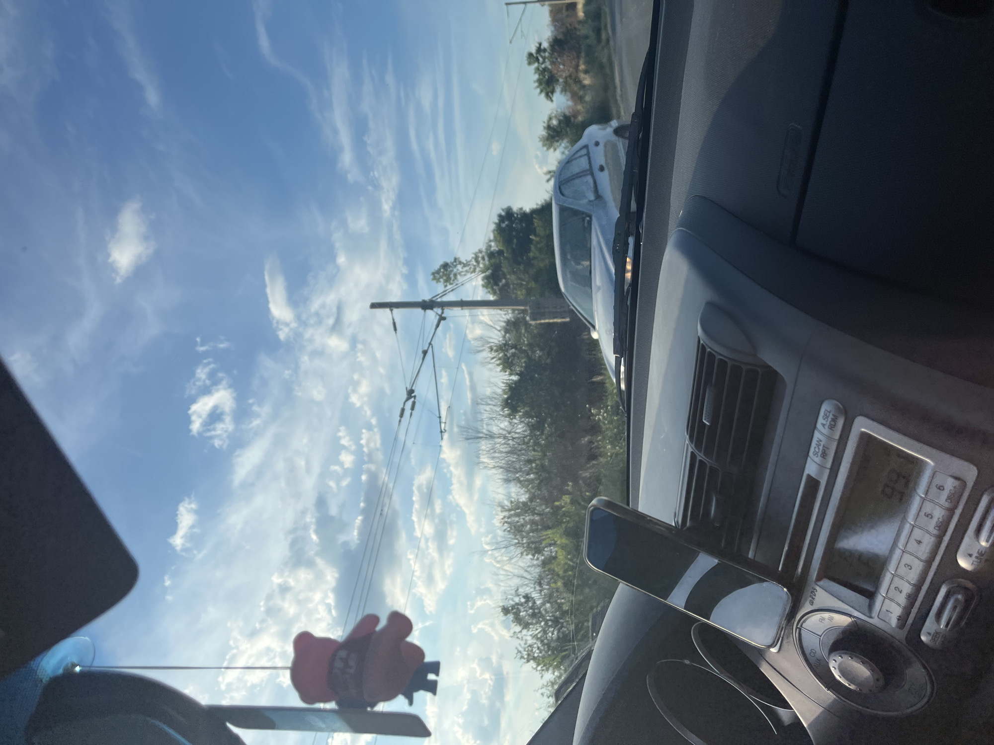 The image is taken from the interior of a car looking out through the windshield. In the foreground, the dashboard is visible with a smartphone mounted on a car mount and the car's radio unit. A pink teddy bear air freshener hangs from the rearview mirror. Outside the car, there is a silver sedan driving in the same direction. The background shows a blue sky with scattered clouds, trees, and utility poles with overhead wires. The scene appears to be set on a road in the city of Dalgo Pole, Bulgaria, under a partly cloudy sky.