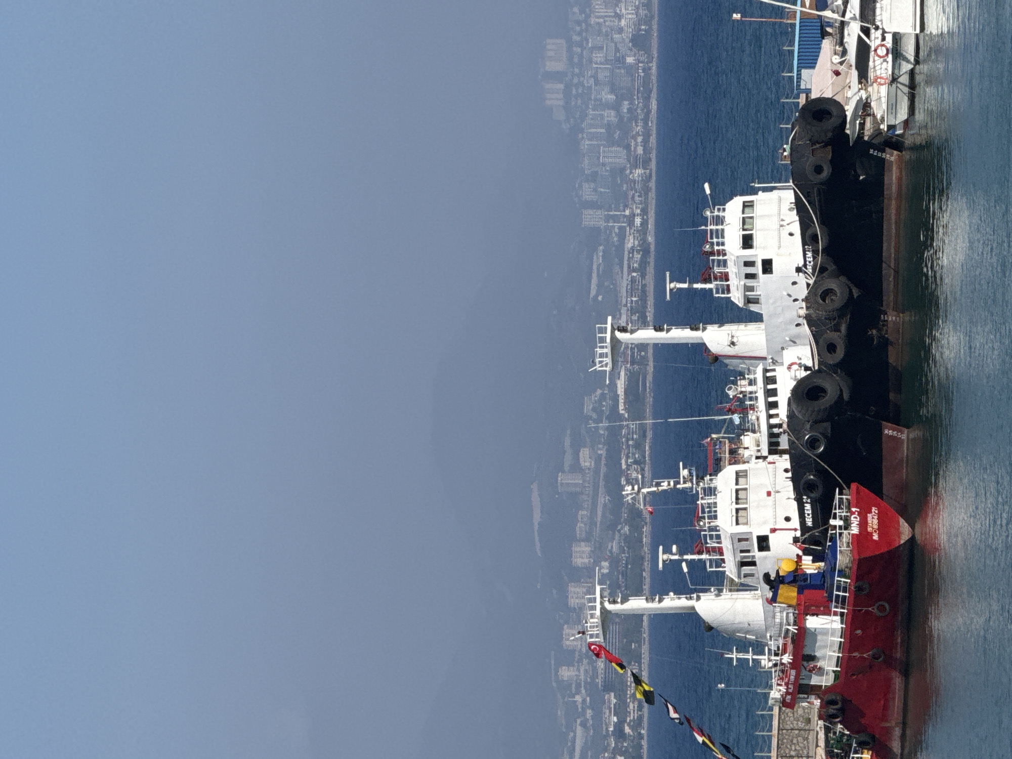 The image shows a view from a ship's deck looking out towards a city on the coastline, with other vessels docked nearby. The city appears densely populated with numerous buildings, suggesting an urban setting. The sea is a deep blue, and the sky is clear and light blue, indicating it is daytime with fair weather.
Several tugboats are visible, prominently featuring a black tugboat with a white superstructure and large black tires along its side. Another tugboat, predominantly red with a white superstructure, is docked next to it. Flags are visible on the vessels, including what appears to be a Turkish flag.
The text "NECEM 22" is visible on the side of the black tugboat. Text on the red tugboat includes "MND-1 ISTANBUL" and "IMO: 0884721".
The scene suggests a working port or harbor, likely in Alanya, Turkey, given the textual clues. The perspective from the ship's deck implies a nautical setting and potentially a moment of rest or observation from a crew member or passenger. There are no people clearly identifiable in the image.