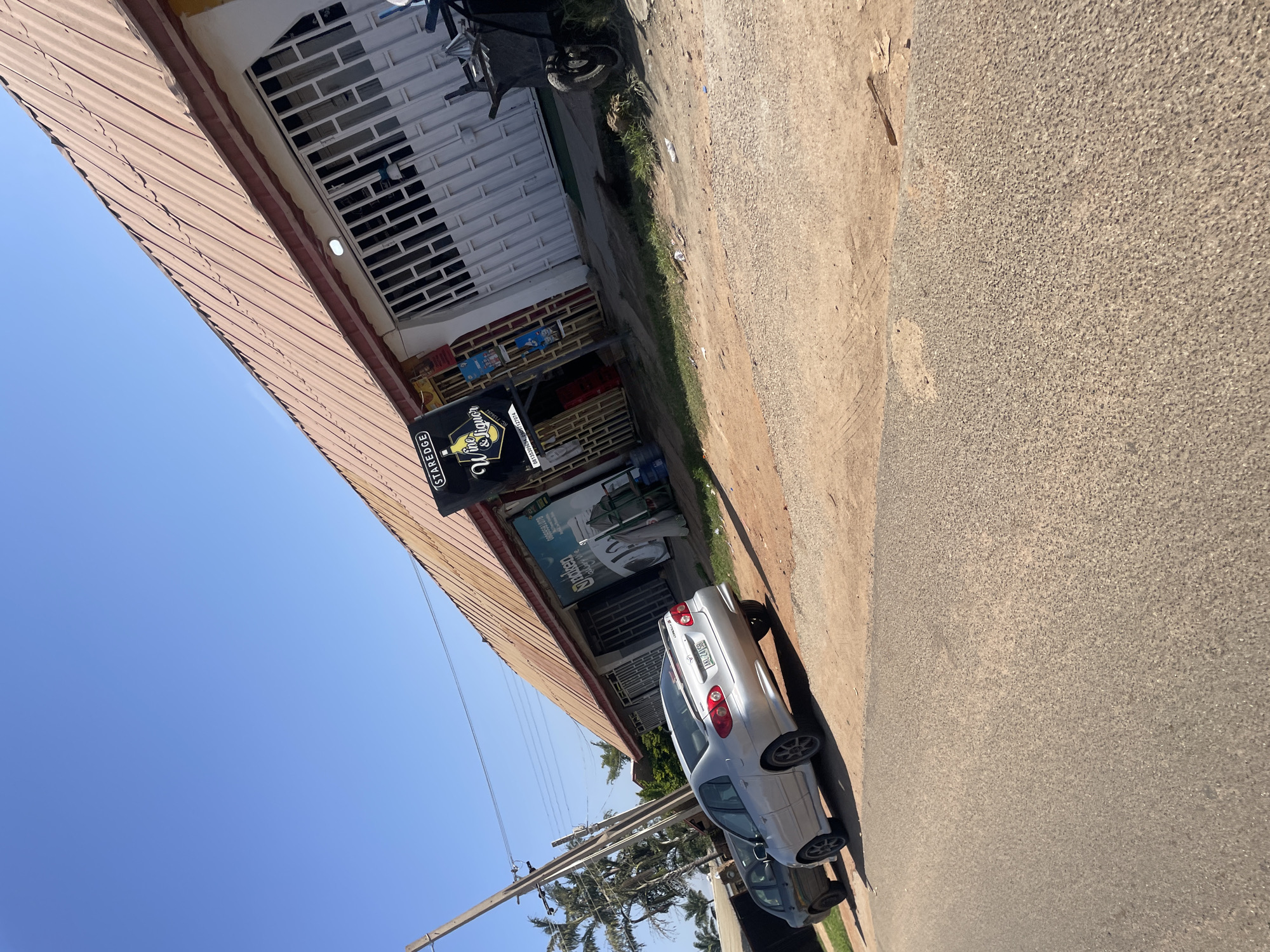 A low-angle, eye-level, medium shot captures a street scene in Gura Top, Nigeria, under a clear blue sky. The focal point is a building with a corrugated metal roof painted a reddish-brown, angled diagonally across the frame from the upper left to the lower right. The building features a white facade with metal security bars covering the windows. A black sign with "STARE DGE Wine & Liquor" in white text is prominently displayed on the left side of the building.
In front of the building, two cars are parked on a dirt and gravel lot. The closest car is a silver sedan with a license plate reading "LSD 241 EH." Behind it is a dark-colored sedan. To the right of the building, a dark gray wheelbarrow is parked on the grass. The street in the foreground is paved with asphalt. Palm trees are visible on the left side of the image, behind a wooden utility pole with wires. The scene appears to be during daylight hours, with bright sunlight casting shadows.