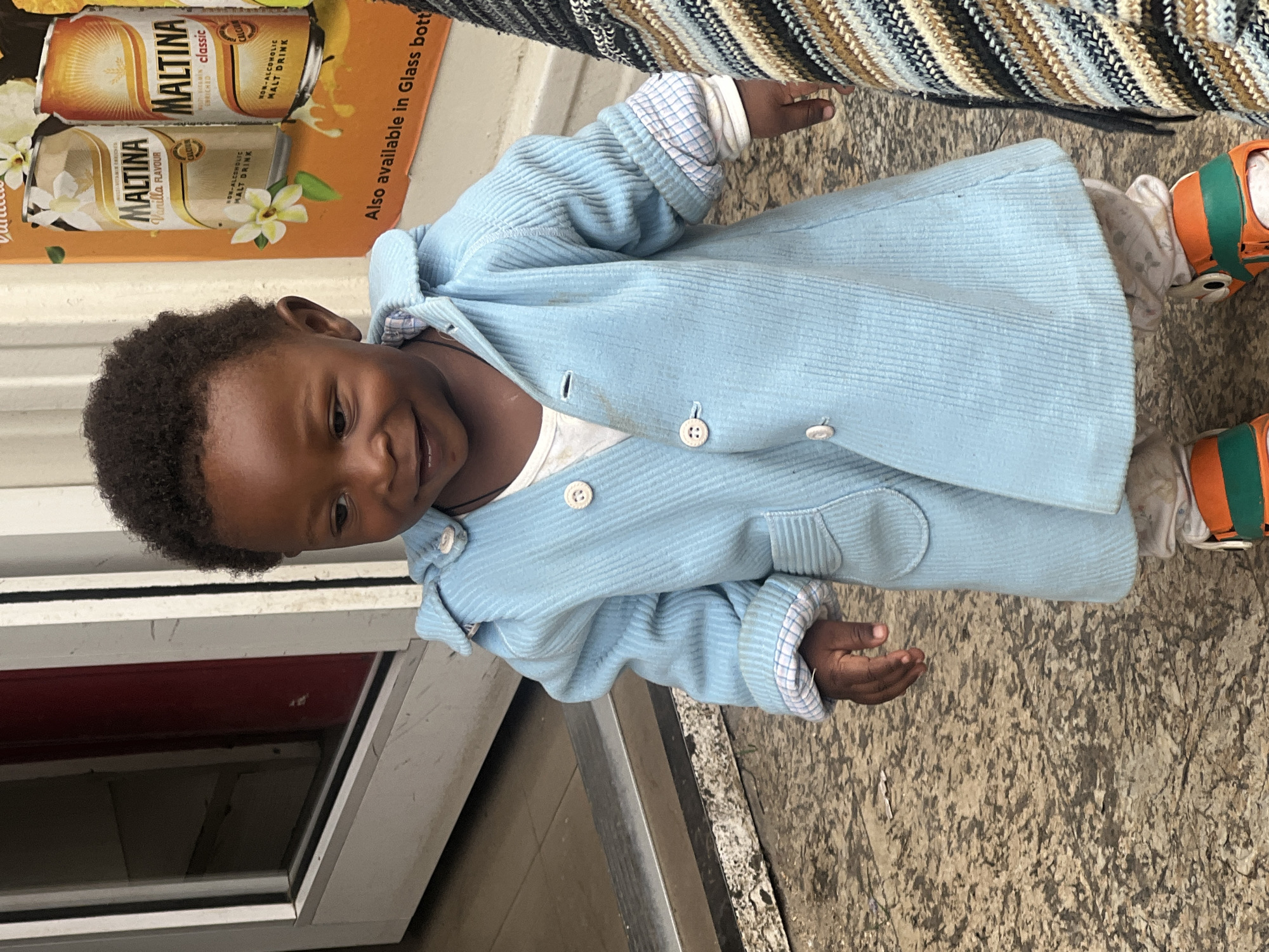 A young, dark-skinned child in a light blue corduroy jacket stands on a textured brown surface, possibly a sidewalk. The child is smiling and looking towards the left of the frame. The child is wearing orange and green shoes. The background includes a window and a display of Maltina beverages, indicating the location is likely a store or vendor stand. Based on the OCR text and the prompt, the location is Gura Top, Nigeria. The child is near the store, possibly interacting with the vendor or just passing by.