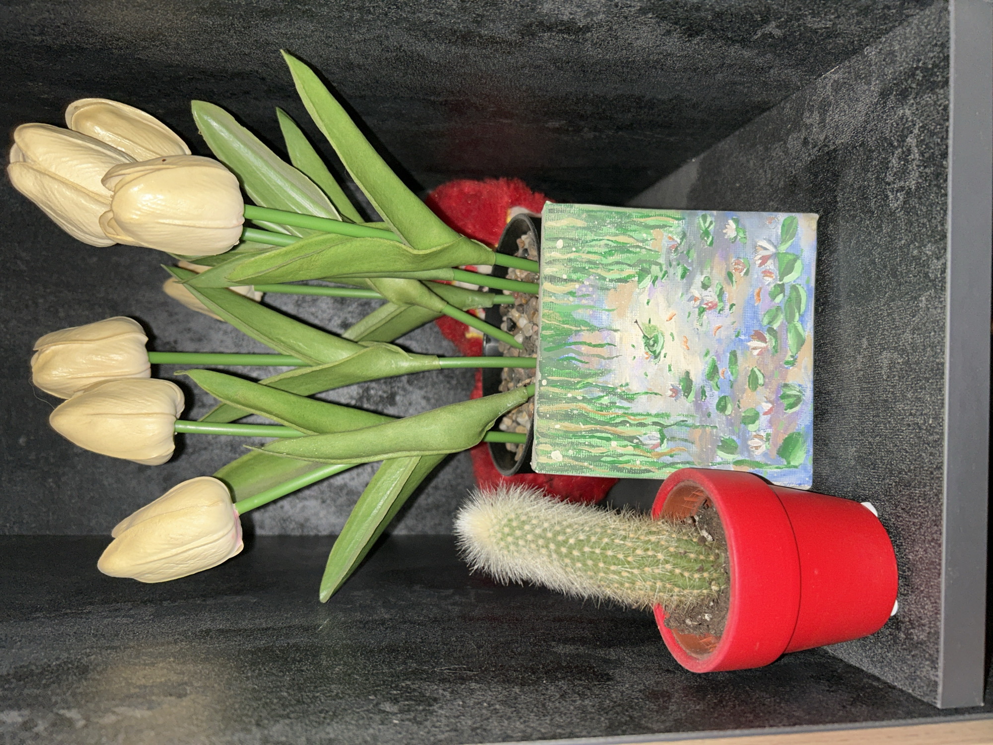 A close-up shot shows a shelf in an urban setting, likely in Sofia, Bulgaria, displaying decorative items. On the left, a small cactus in a bright red pot stands upright, its fuzzy texture visible. To its right, a miniature canvas painting depicts a serene water scene with lily pads and fish. Behind these, a cluster of artificial cream-colored tulips with green stems and leaves are arranged in a pot. A soft red object, possibly a plush toy, peeks out from behind the tulips. The shelf itself is dark gray and textured, contrasting with the darker, rough-textured wall behind it. The overall scene suggests a home or office interior, showcasing a collection of indoor plants and decorative art.