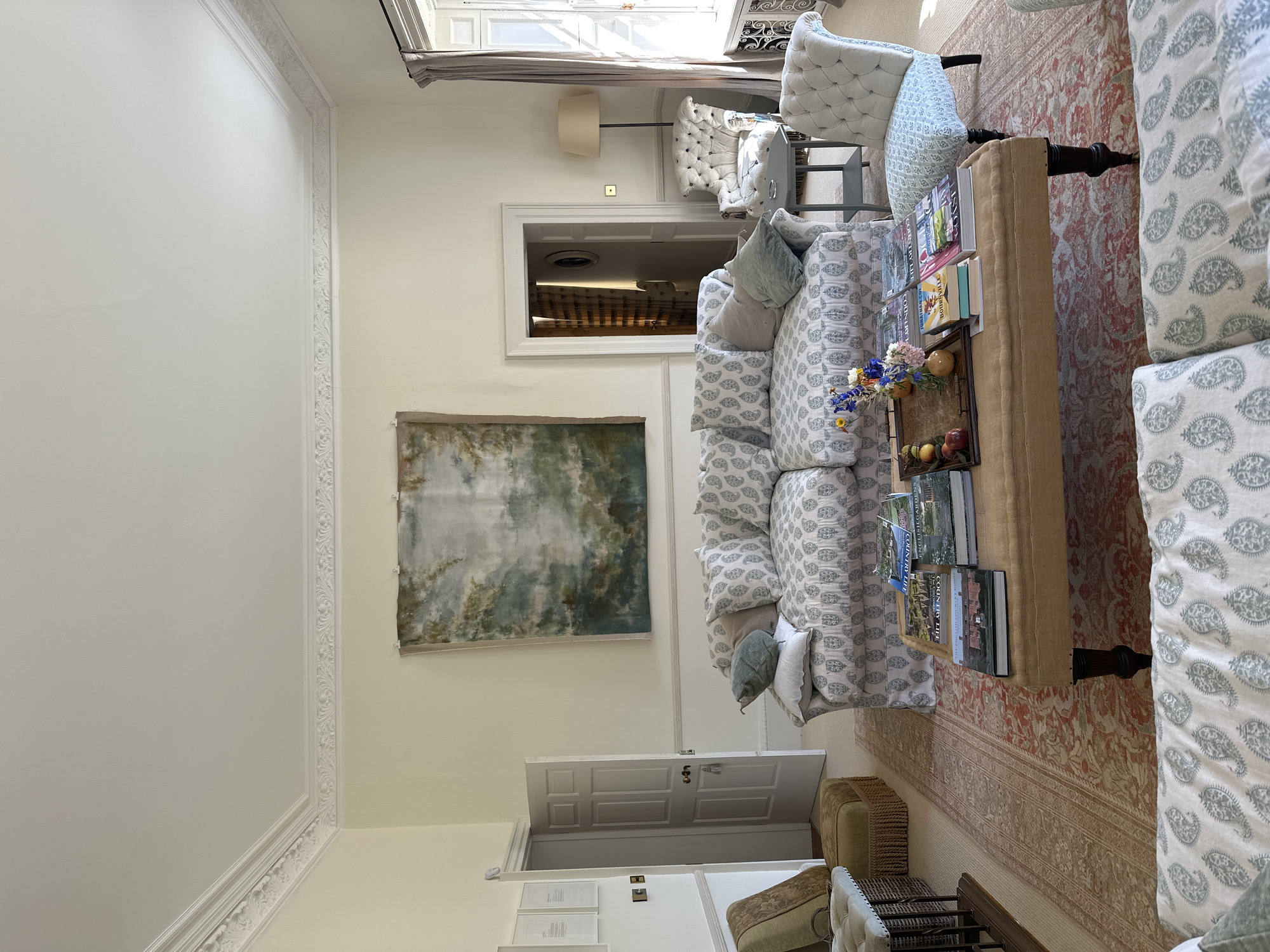 The image shows an interior living room space. The walls are painted in a neutral tone, and the ceiling has decorative molding. A large abstract painting adorns one wall. Furniture includes sofas and chairs upholstered in a paisley pattern, a light-colored carpet with an oriental design, and a coffee table. Books and decorative items are placed on the coffee table. A doorway leads to another room. The location is in the city of Unknown, Unknown.