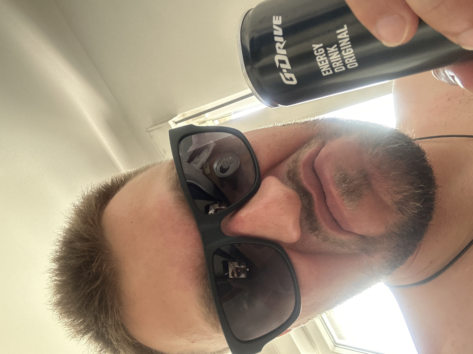 The image shows a close-up, low-angle shot of a person holding a can of "G-DRIVE ENERGY DRINK ORIGINAL." The person is wearing dark sunglasses that reflect a small image, possibly of themselves. The background is a plain, light-colored wall or ceiling, with a window or light source visible in the upper right corner, suggesting it's daytime. The lighting creates a bright, slightly blown-out effect in parts of the image. The person has short, light brown hair and stubble on their chin. The scene appears to be indoors, possibly in a home or office, given the indoor lighting and wall texture. The overall impression is informal, with the subject looking directly towards the camera. The visible text on the can clearly reads "G-DRIVE ENERGY DRINK ORIGINAL." The context of Zvezdara Urban Municipality, Serbia, does not provide any specific visual cues in the image itself.
