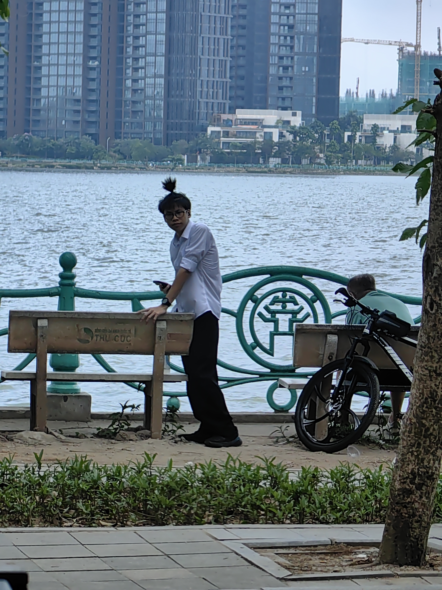 The image shows a man with a ponytail wearing glasses and a white shirt standing beside a bench along a waterfront promenade. He appears to be holding a phone. In the background, across the water, are tall apartment buildings and a construction site with a crane. A green metal railing separates the walkway from the water. To the right, there is another person seated on a bench, partially obscured by a bicycle and a tree trunk. The benches have "THU CUC" printed on them. The location is likely in the city of Hà Nội, Vietnam, given the Vietnamese writing on the bench.