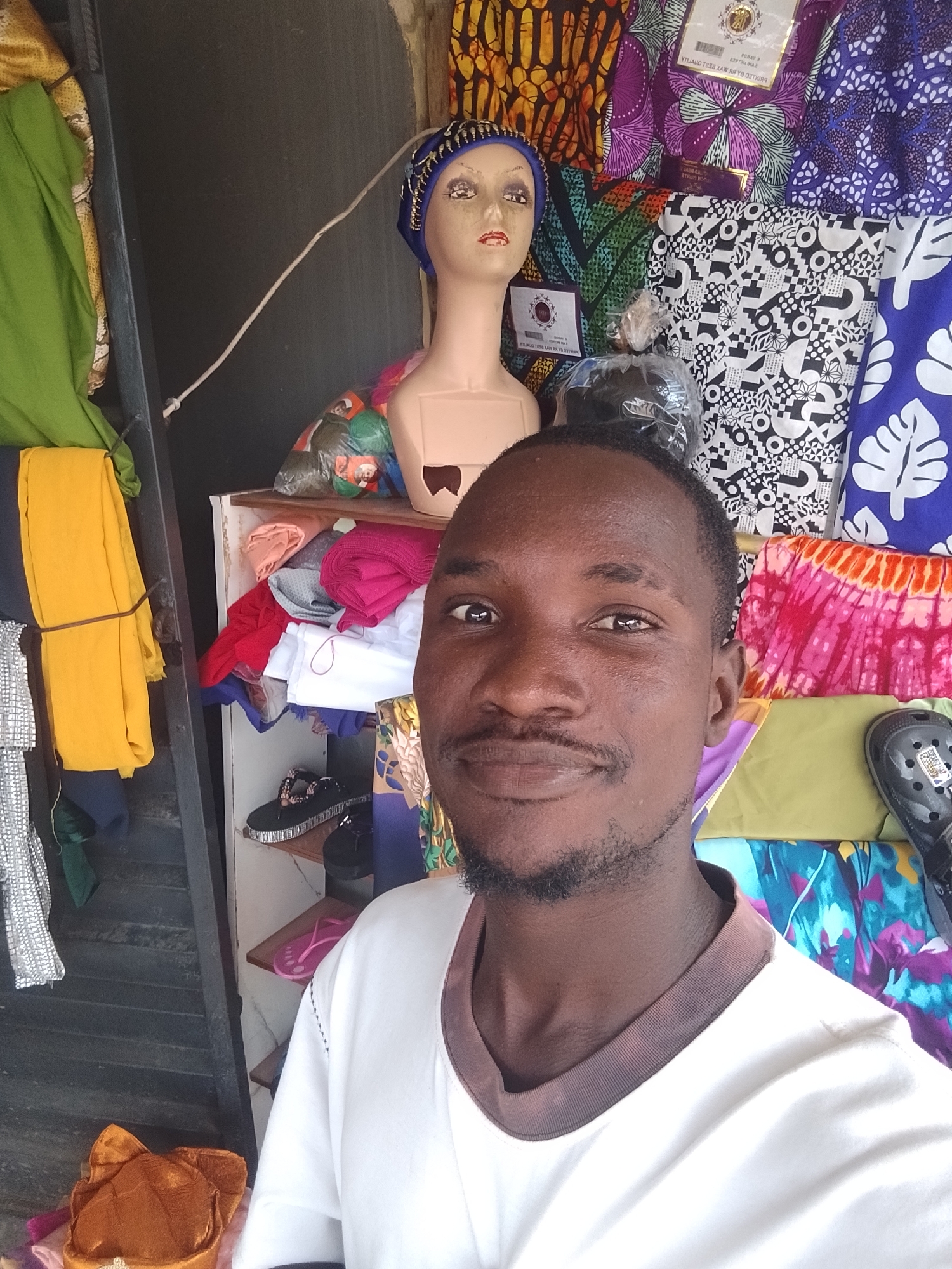 A medium shot shows a man with dark skin, a mustache, and a goatee, wearing a white shirt with a brown collar. He is standing in front of a stall displaying various colorful fabrics, a mannequin head wearing a blue and gold headscarf, and shelves stocked with folded fabrics, a pair of black embellished slides, and pink flip-flops. In the foreground, to the left, is a stack of folded fabrics, including a bright yellow one tied with string. A brown and gold cap sits on top of a pile of fabrics in the bottom left corner. The stall appears to be located in Ado, Nigeria, suggested by the display of vibrant fabrics commonly seen in West African markets. The lighting is bright, indicating an outdoor or well-lit indoor setting.