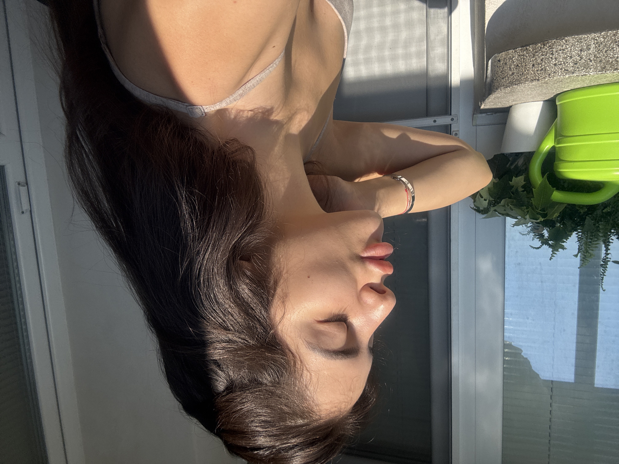 A young woman is captured in profile, with her eyes closed and lips slightly pursed, as sunlight illuminates her face and hair. She is wearing a light-colored tank top and a bracelet on her left wrist. Her hair is long and dark brown, falling over her shoulders. To her left, a bright green watering can sits on a stone ledge next to a potted plant. Behind her, a window with blinds and a door with a screen are visible. The scene appears to be an outdoor balcony or patio, with shadows indicating a sunny day. The specific location in Plovdiv, Bulgaria, is not discernible from the image itself.