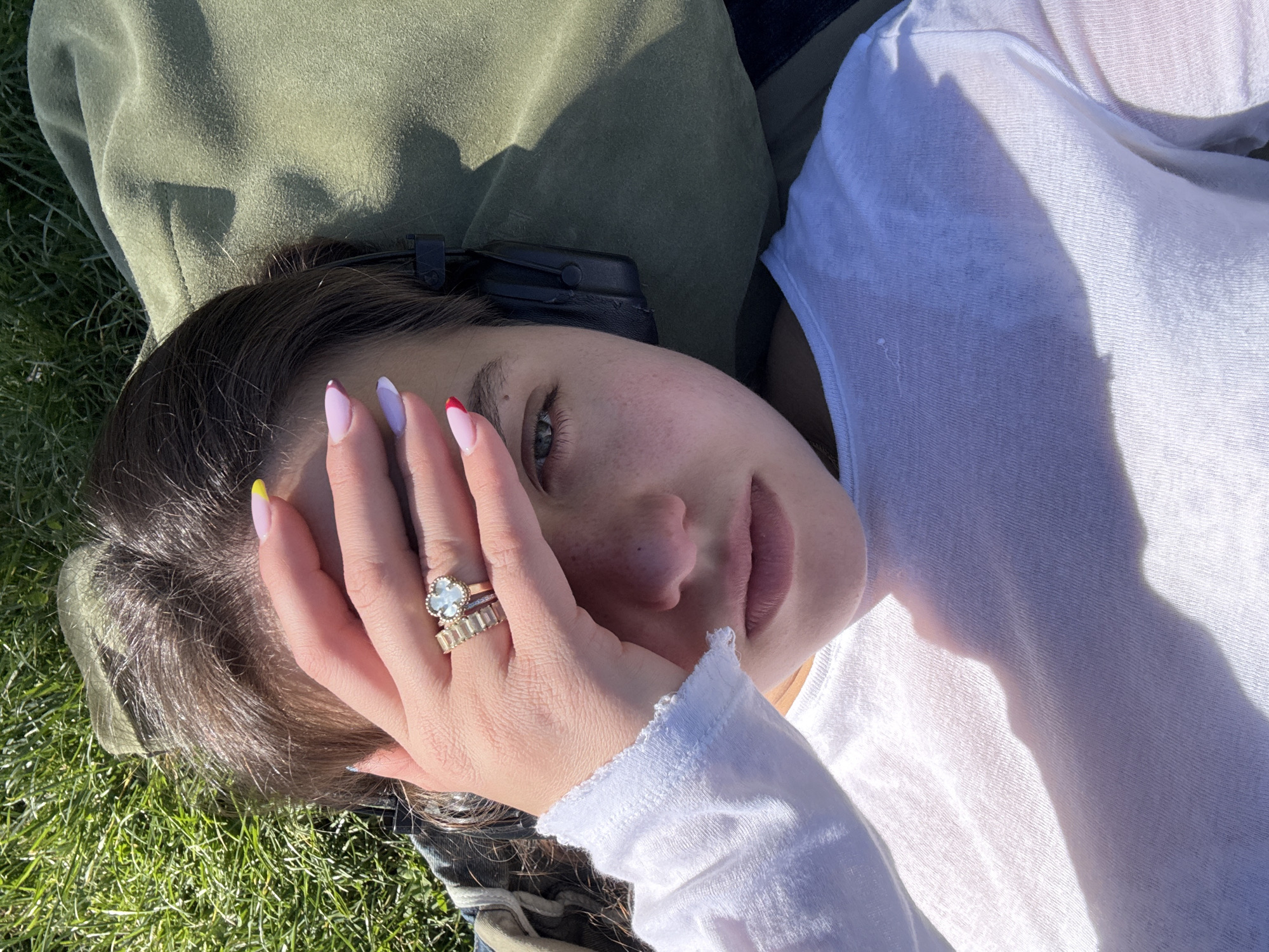 A fair-skinned young woman is lying on grass, her head resting on a green jacket or pillow. She is wearing a white long-sleeved shirt. Her right hand is raised near her face, displaying several rings on her fingers and a manicure with various colored tips (yellow, purple, red, and brown). The woman has brown hair, fair skin with freckles, and light-colored eyes. A black object, possibly headphones, is visible behind her head. The background includes grass and part of a blue garment, likely denim jeans. The image is taken outdoors in natural lighting. There is no information that specifies the location in the city of Milan, Italy.