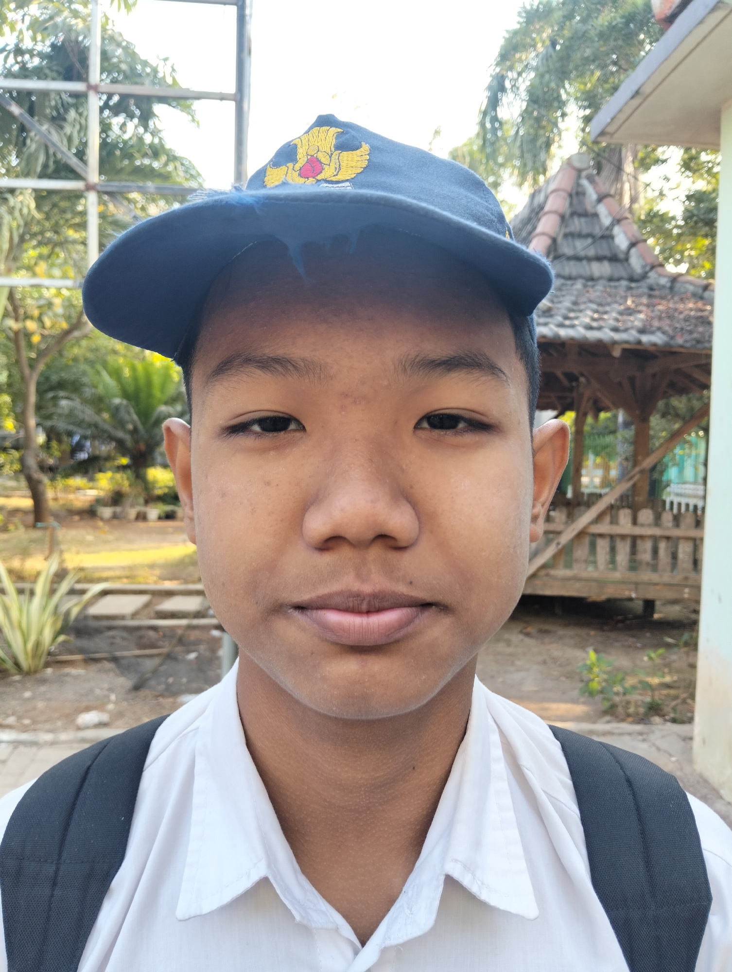 A young Indonesian boy stands outdoors, facing the camera. He wears a dark blue baseball cap with a yellow emblem, a white collared shirt, and a black backpack. The background shows a tiled roof structure, trees, and some foliage, suggesting an outdoor setting likely in a semi-rural or suburban area. The light suggests it is daytime.