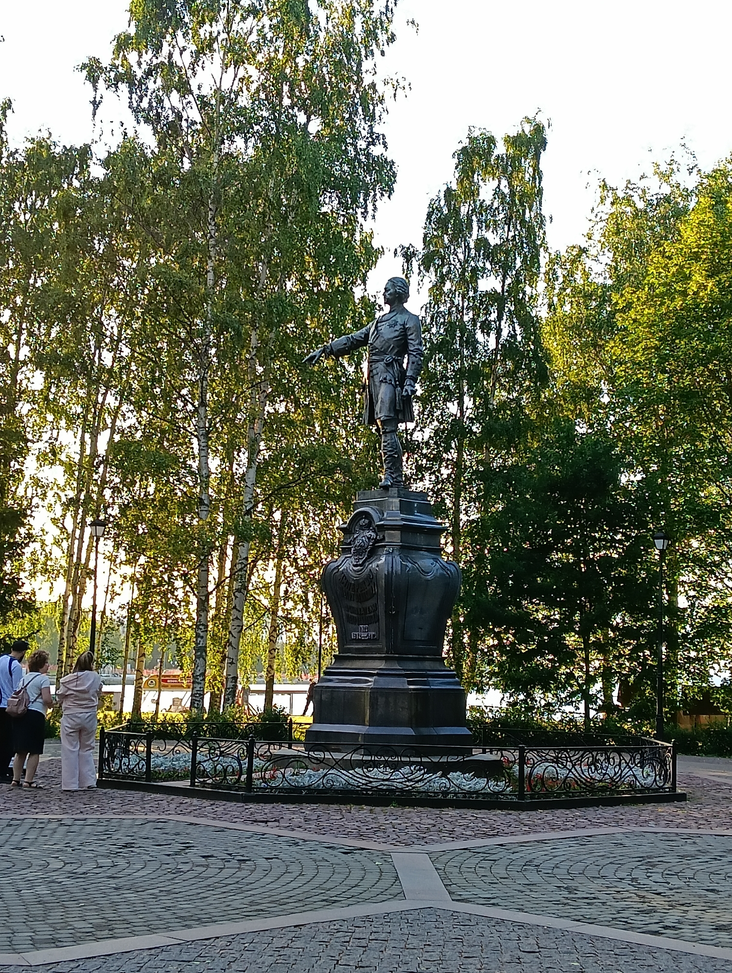 A bronze statue of Peter the Great stands on a stone pedestal in a park in Petrozavodsk, Russia. The statue depicts Peter the Great in military attire, pointing to the right. He is wearing a sash and a sword. The pedestal is adorned with decorative carvings and a plaque with inscriptions. A decorative black metal fence encloses the area around the pedestal.
In the foreground, a paved area made of interlocking stones forms a circular pattern. Several people are visible in the background. To the left of the statue, three individuals are standing. One person, wearing a white shirt and dark pants, is looking towards the statue. Another person, in a light pink tracksuit, is facing away from the camera, looking towards a body of water. A third person, wearing a dark skirt and a white top, is also facing away.
Behind the statue, a line of birch trees with green leaves creates a natural backdrop. The sun is shining, casting a warm light on the scene. In the distance, a boat with a red roof is visible on the water. The overall environment suggests a pleasant outdoor setting, likely a public square or park within the city. The statue is a prominent monument, suggesting historical or cultural significance.