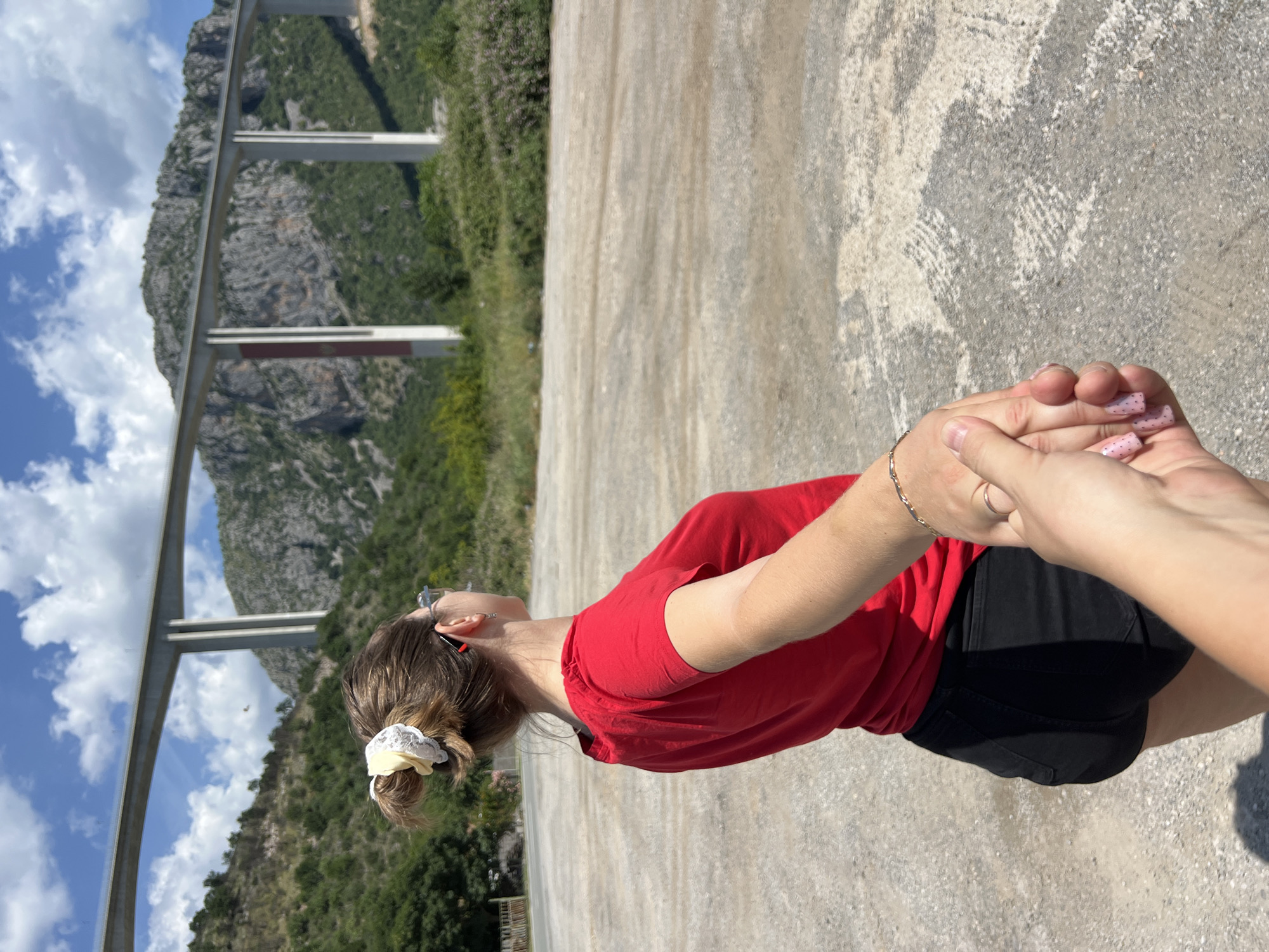A photograph taken in the city of Bioče, Montenegro, shows a person with medium-length brown hair pulled back with a white scrunchie. The person is wearing a red t-shirt and black shorts. Their right hand is being held by another person, whose nails are painted light pink with small black dots. Behind the individuals, a concrete road and bridge are seen against a backdrop of a mountain landscape. The sky above is blue with white clouds.