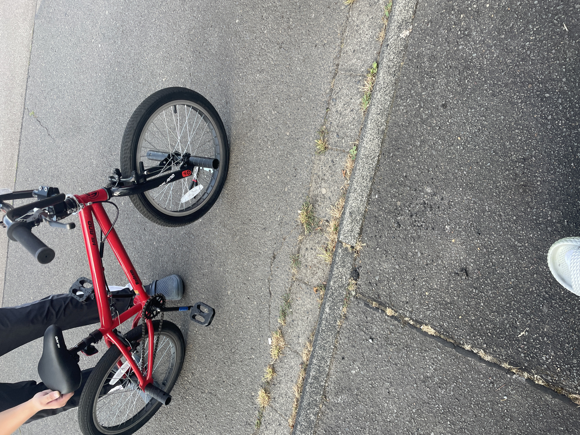 A red HILANO "Redeem" BMX bicycle is positioned on an asphalt surface. The bicycle is in the center of the image, facing slightly to the right. It has black handlebars, a black seat, and black pedals. The front tire appears to be slightly deflated. A person, partially visible from the waist down, is standing to the left of the bicycle, holding the seat with their left hand. They are wearing black pants with white stripes and grey Crocs-style shoes. The background consists of more asphalt, with some cracks and sparse vegetation growing through them, suggesting an outdoor, possibly urban or suburban, environment. The scene does not provide specific landmarks to confirm its location in Wiehl, Germany.