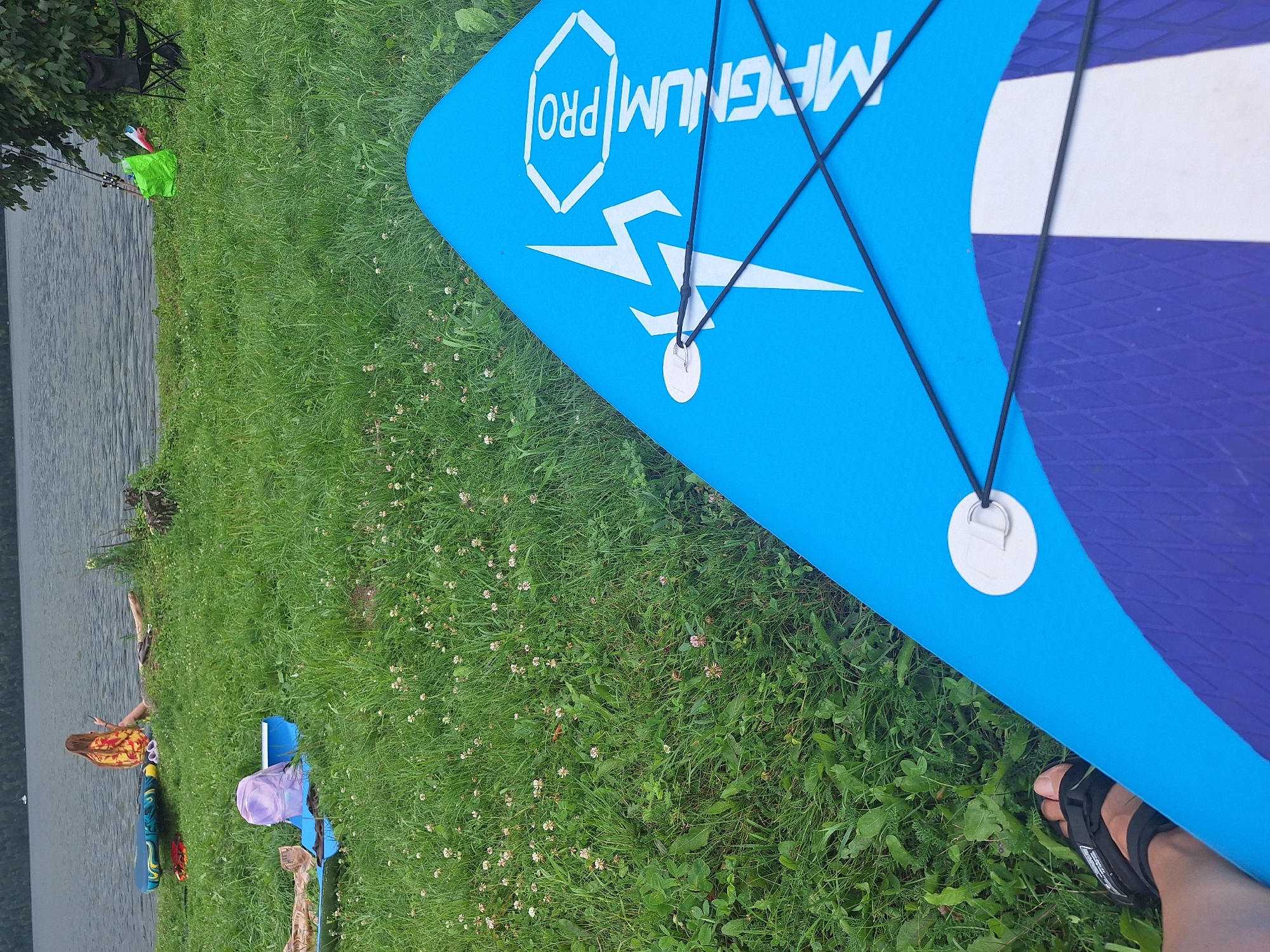 The image depicts a natural scene on the shore of a body of water, likely a lake, with a foreground dominated by a blue paddleboard. A person's sandaled foot is visible in the lower left corner, suggesting they are either on the paddleboard or standing near it. In the middle ground, several other objects are scattered on the grass, including a paddleboard, a blue mat with a purple backpack on it, and a partially visible tan object. Further back, a young woman with long hair, wearing a colorful sarong, is seated by the water's edge, holding a phone. Near her are two white sandals and another paddleboard. The background reveals a body of water and a tree line, with fishing rods leaning against a tree and a green bag and a folding chair nearby. The environment is grassy and appears to be a public recreational area. The specific location of Atamanovka, Russia, is not explicitly identifiable from the visual information in the image alone, but the general setting suggests a lakeside or riverbank.