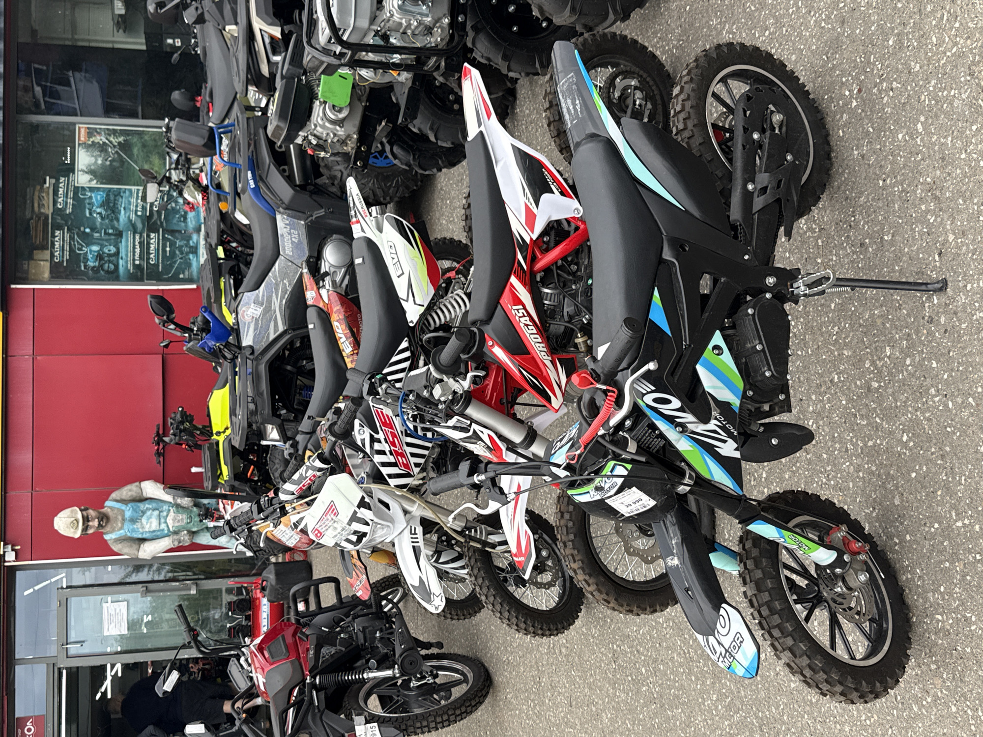 The image depicts an outdoor scene in front of a building with a red facade, likely a retail establishment. The location is identified as Votkinsk, Russia. In the foreground, a row of dirt bikes and ATVs is parked on an asphalt surface. The vehicles are of various colors and designs, with some displaying branding such as "BSE" and "KAVO MOTOR". One dirt bike in the very front has a price tag of "29 990" affixed to it. Behind the vehicles, a life-size statue of a construction worker in blue overalls and a hard hat stands near the entrance of the building. The entrance itself is a glass door with a visible sign. Further back, several ATVs are parked. The overall impression is that of a dealership or outdoor display area for motorized vehicles.