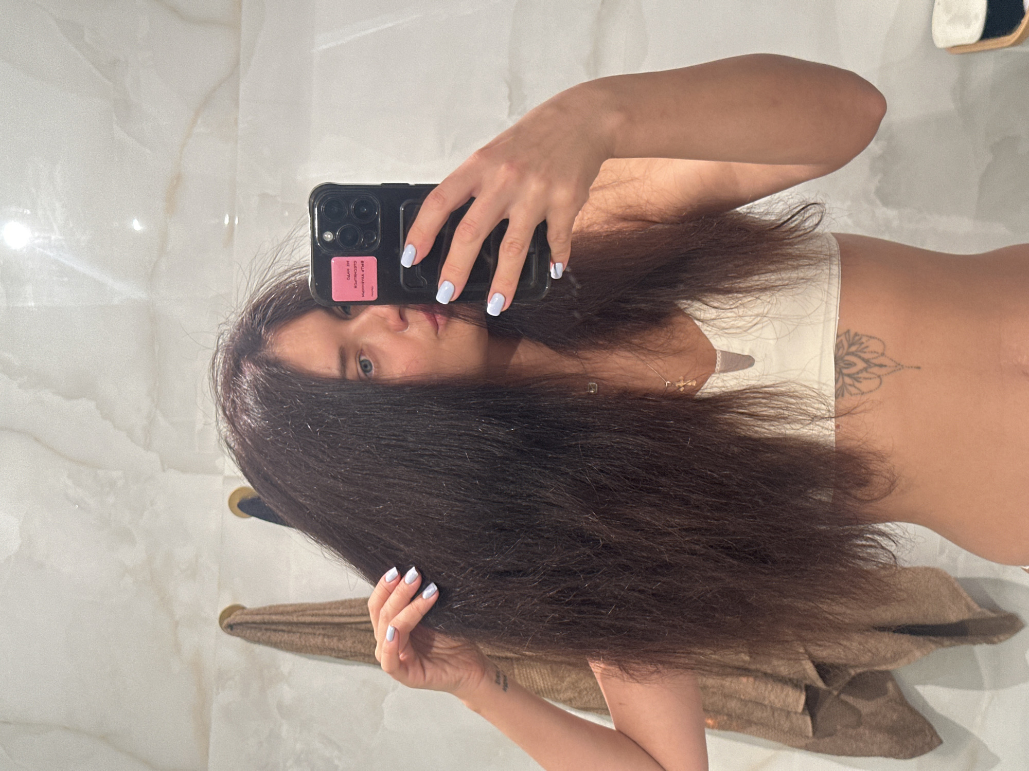 A young woman is captured in a selfie taken in a mirror, possibly in a bathroom or dressing area, within an indoor setting in Moscow, Russia. She is shown from the chest up, with her long, dark brown hair flowing down and covering parts of her body. Her right arm is raised, holding a black smartphone with multiple camera lenses and a pink sticker on the back. Her fingernails are painted a light blue or lavender shade. Her left hand is visible further down, holding a section of her hair.
She wears a light-colored top, possibly a bralette, and a delicate necklace with a small cross pendant. A mandala-like tattoo is visible on her right side/ribcage area. The background consists of smooth, reflective, light-colored walls, likely polished stone or marble, creating a clean aesthetic. A brown towel is draped over a rail or hook in the background. The woman looks directly at the camera with a neutral expression.
Visible text on the pink phone sticker includes Cyrillic characters: "ВЫШЛА УДАЧНОЙ СЛЕСЬМИСЯ НЕ НУТО".