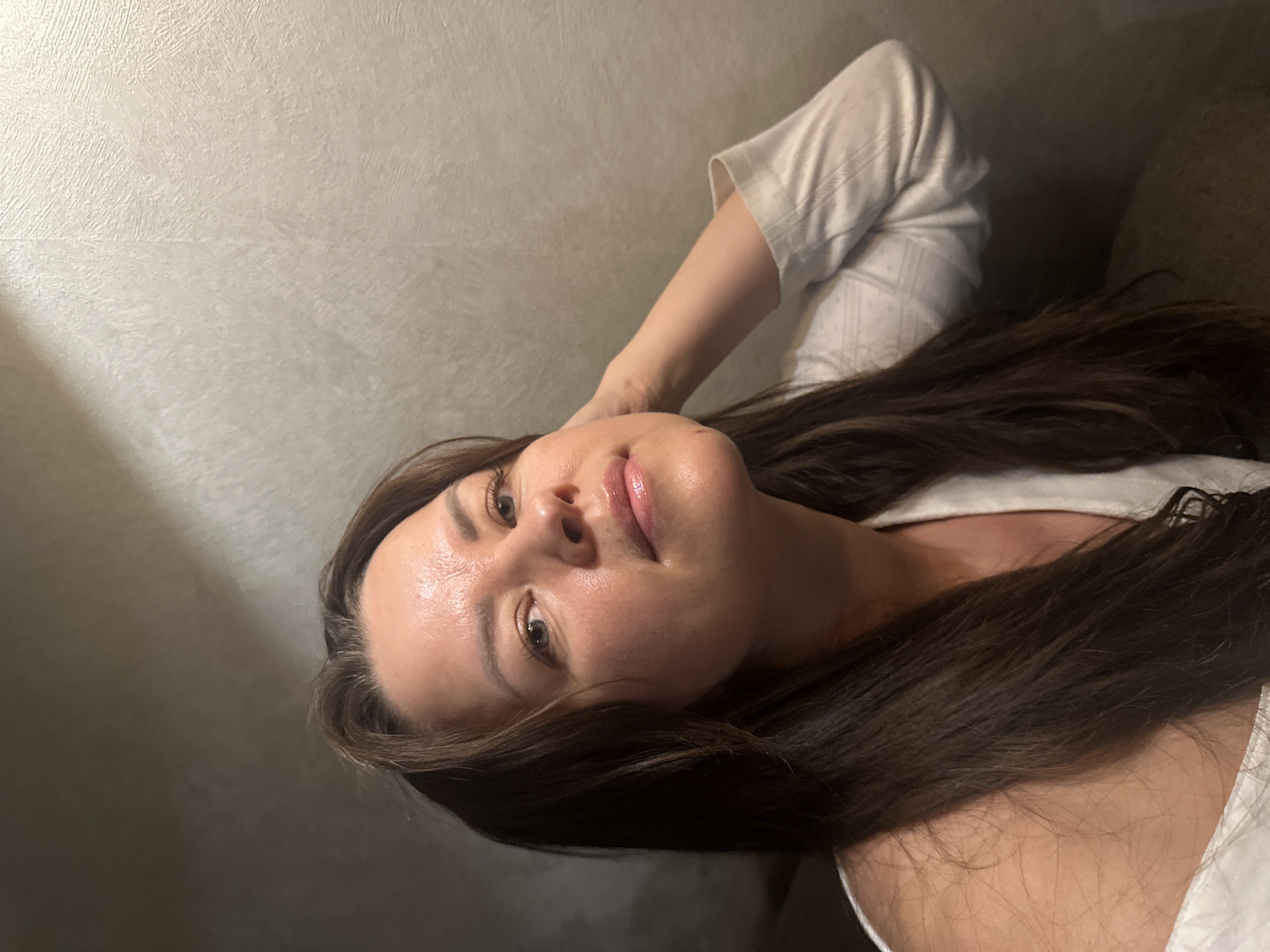 This close-up portrait, possibly a selfie, features a woman with long, dark brown hair and light-colored eyes, likely green or grey. She is depicted in a relaxed pose, seemingly lying down, with her head slightly tilted and one arm casually placed behind her head, allowing her hair to fan out. Her facial expression is serene and contemplative, with a subtle smile and direct gaze towards the camera. Her skin appears smooth with a slight sheen, and her lips have a glossy finish. She is wearing a light-colored, possibly white or off-white, top with a visible textured pattern on the sleeve. The background is a plain, light grey or beige textured wall, suggesting an indoor setting. Soft, diffused lighting illuminates her face, hinting at an indoor environment, possibly evening or a room with indirect natural light. There are no other prominent objects or activities. No text is visible in the image. Although the location context is Chaikovsky, Russia, the image itself does not contain specific visual cues to identify this location. The overall atmosphere conveyed is calm and peaceful.