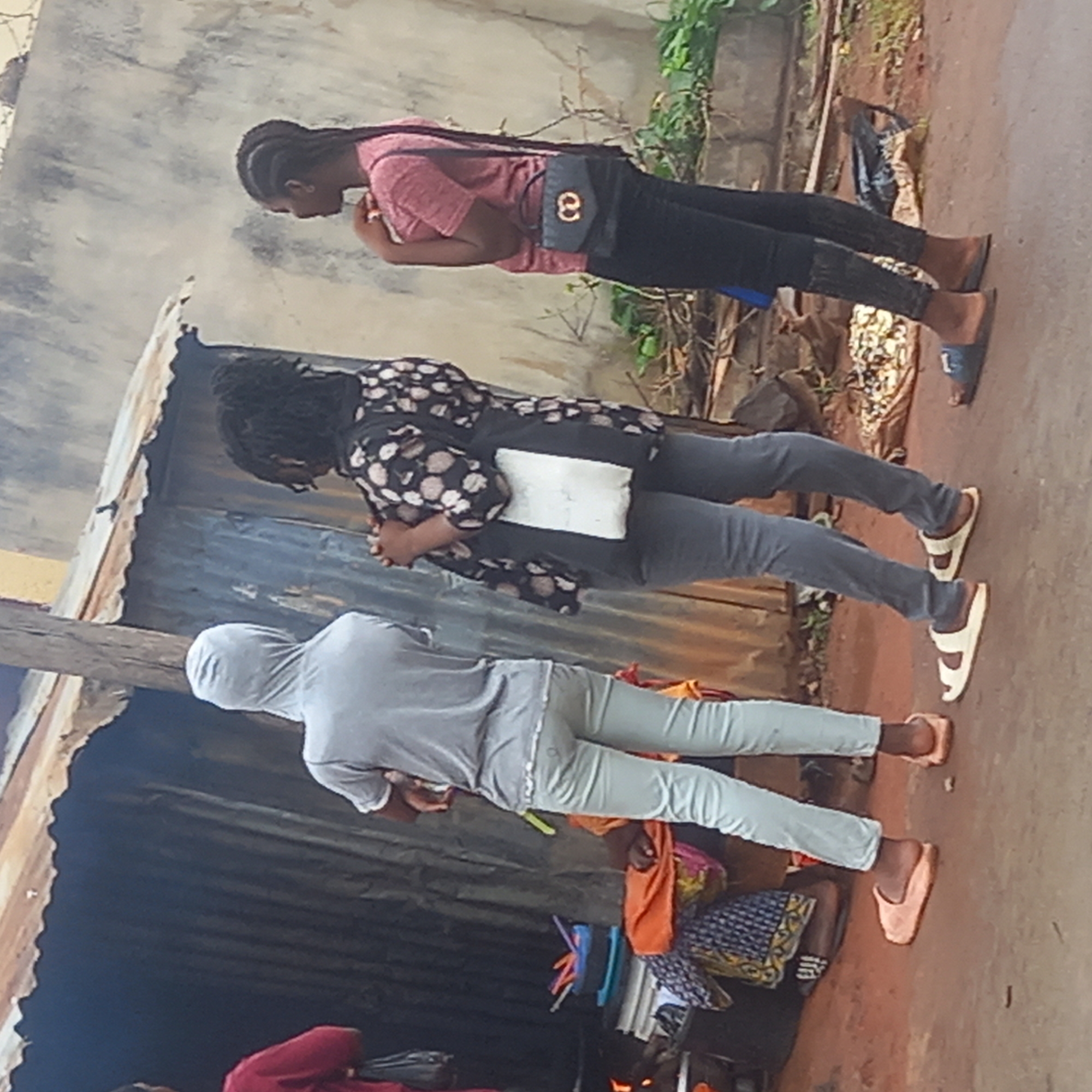 Three individuals stand aligned on a paved surface in Barden Yana, Nigeria. The person farthest from the camera is wearing a gray hooded sweatshirt and light-colored pants. The middle person has on a black and white patterned shirt, gray pants, and white sandals. The person closest to the camera is wearing a pink shirt, black pants, and blue sandals. All three have their heads bowed and hands clasped in front. Adjacent to the paved area is a corrugated metal structure, likely a building or shed. There is a pile of various items beneath the structure near the person in gray. Vegetation is seen behind the person in pink.