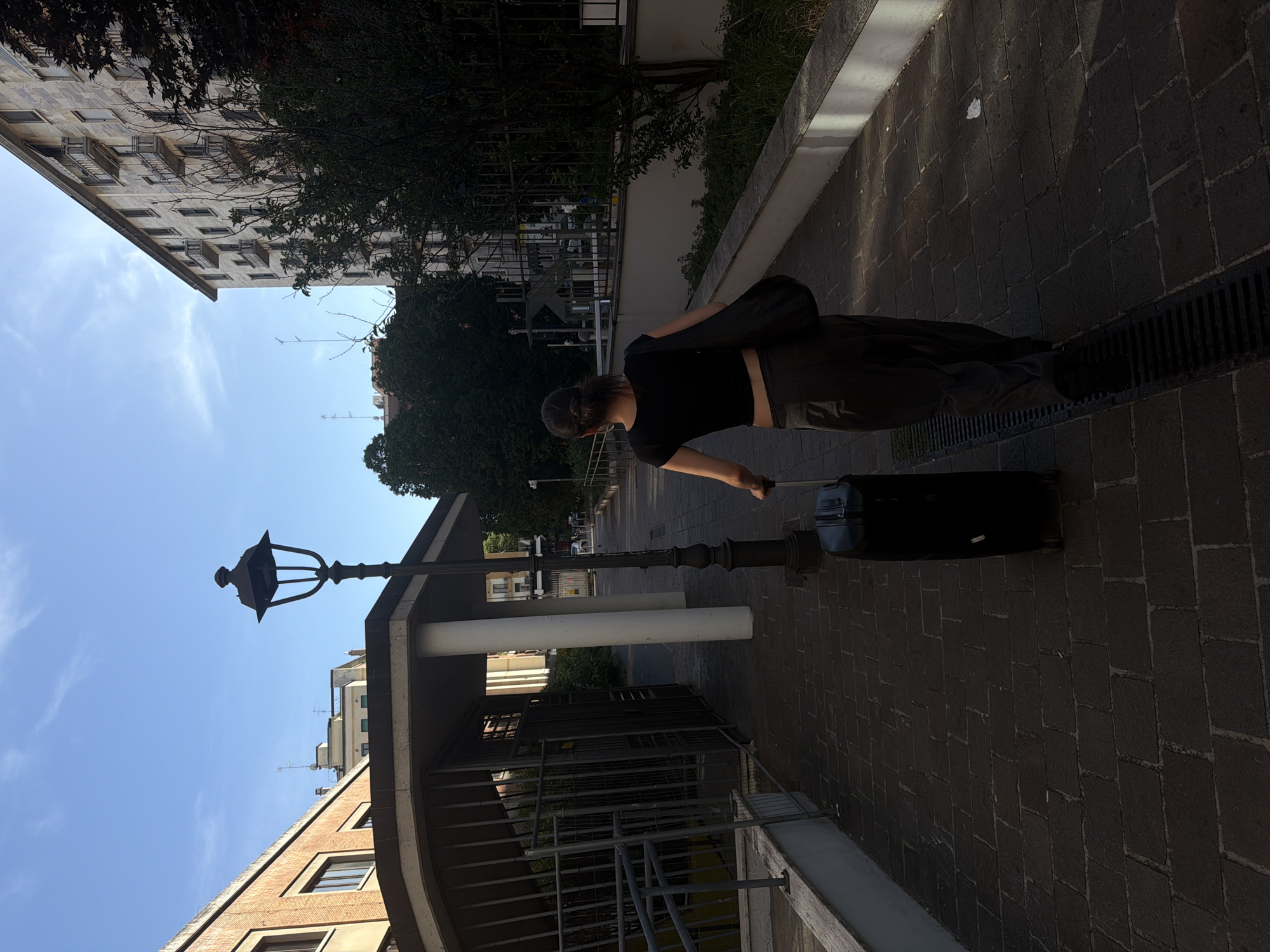 A young woman with dark hair pulled back in a ponytail walks away from the camera, pulling a black wheeled suitcase behind her. She is wearing a black crop top and dark, loose-fitting pants, and has a black shoulder bag slung across her body. The woman is walking on a brick pathway that runs alongside a white wall and a drain grate. A black lamppost stands to her left. In the background, there are buildings with balconies and trees, as well as a fenced-off area with a ramp. The scene is set in a city environment, likely a residential or urban area in Milan, Italy, given the architectural style and urban setting. The lighting suggests it is late afternoon or early morning.