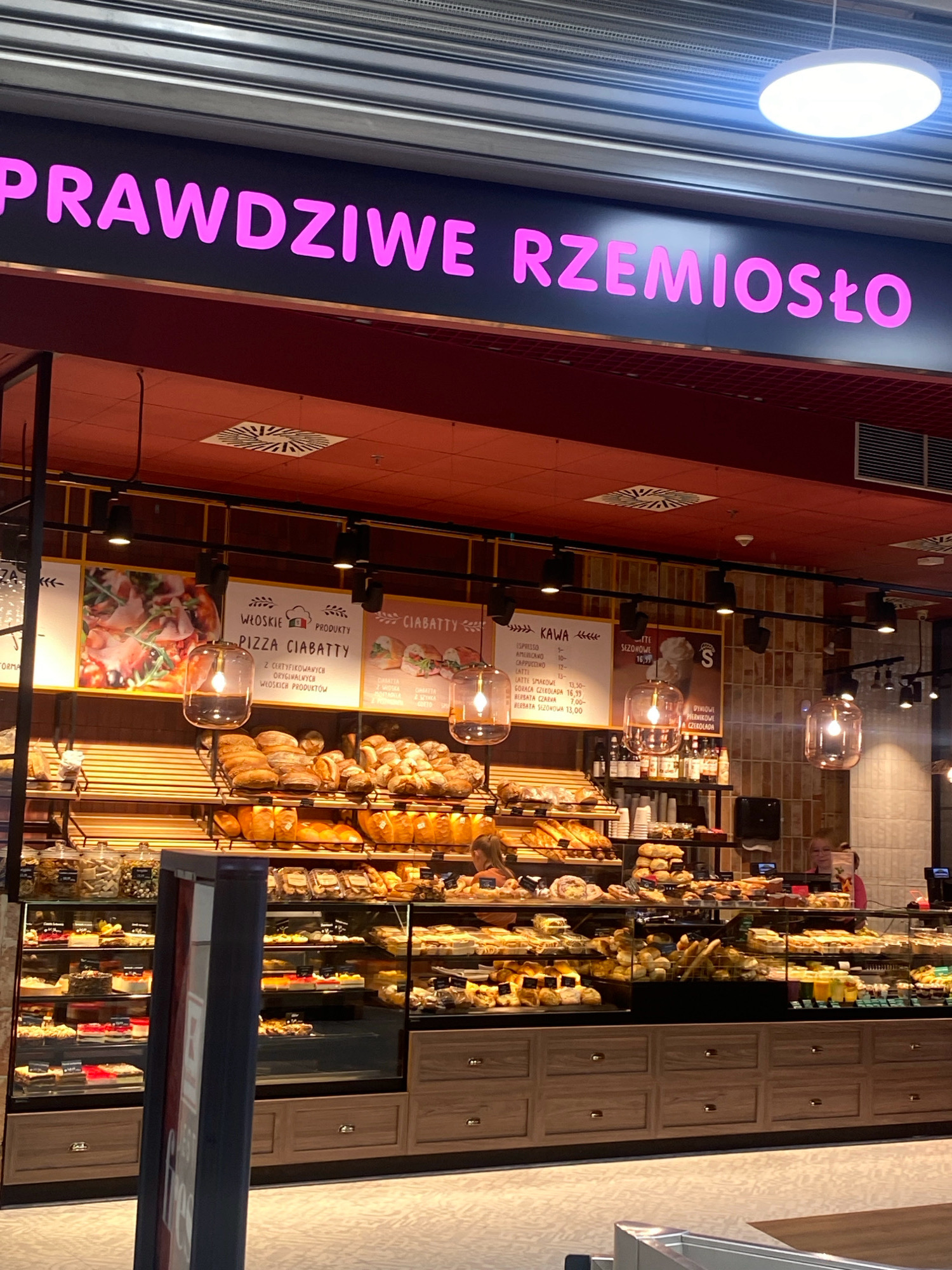 The image shows the interior of a bakery with a prominent neon sign that reads "PRAWDZIWE RZEMIOSŁO" in pink lettering. The bakery features a wide selection of bread and pastries displayed on shelves and in refrigerated cases. Several people are visible, including a woman behind the counter and customers browsing the goods. The overall atmosphere suggests a modern and well-stocked establishment. Based on the presence of Polish text and the specific design elements typical of such shops in Poland, this bakery is likely located in Nowy Sącz, Poland.
