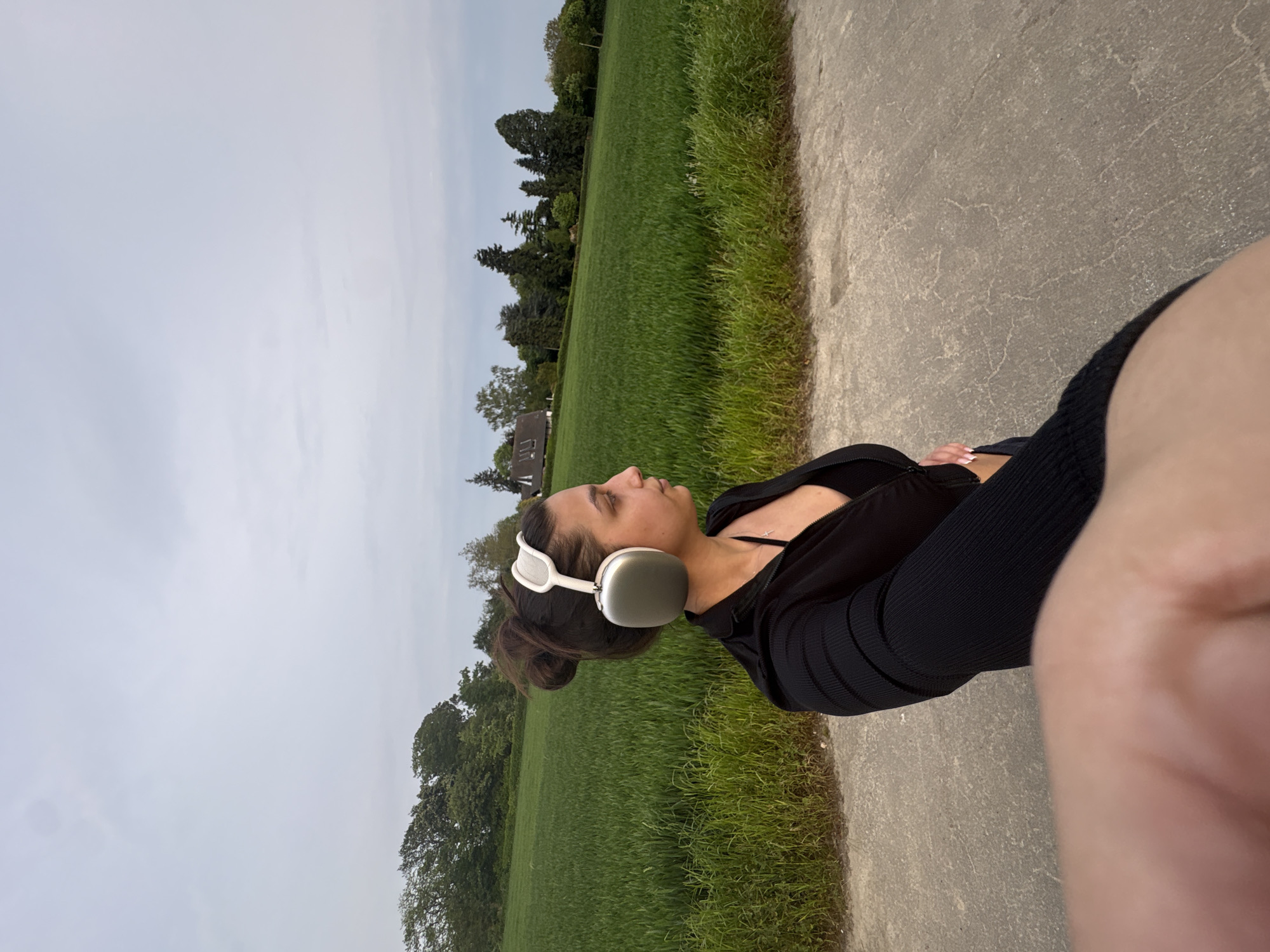 A woman is standing on a paved path next to a grassy field. She is wearing a black top and leggings and has white headphones on. The field is green and stretches into the distance, where there are trees and a building. The sky is overcast. The location is likely in Mönchengladbach, Germany, based on the user's request. The image appears to be a selfie, with part of the person's arm and hand visible in the foreground.