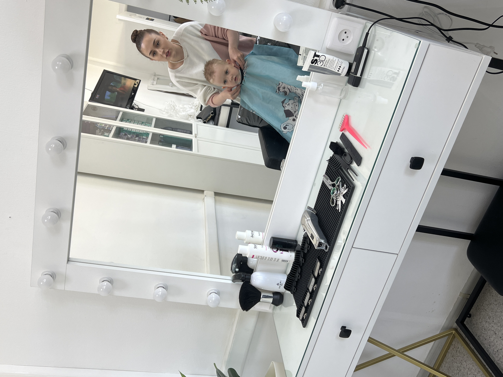 A mirrored vanity desk in a room is seen. The room contains a young woman and a small child. The woman is standing behind the child and is wearing a white sweater and a strand of pearls. The child is wearing a blue cape with Dalmatians printed on it. The cape is clipped around the child's neck. The vanity desk has a mirrored surface with light bulbs around the frame. On the desk are various hair-styling tools, including a comb, scissors, clippers, and bottles of product. A television and display shelves are visible in the mirror's reflection. The location is Chaikovsky, Russia. The woman appears to be giving the child a haircut.