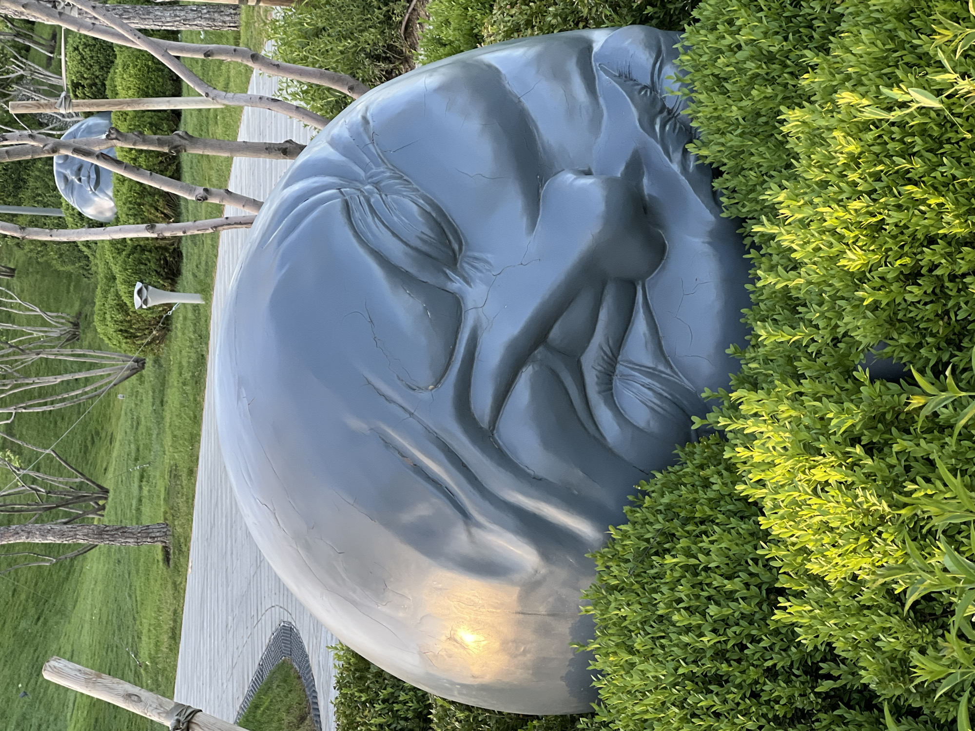A large, gray, spherical sculpture of a human face lies on its side in a park-like setting. The sculpture is cracked and appears weathered. Lush green bushes partially obscure the right side of the face. A wooden boardwalk extends behind the sculpture, leading to a grassy area with sparse trees. There are similar smaller sculptures in the distance. The location is Krasnodar, Russia.