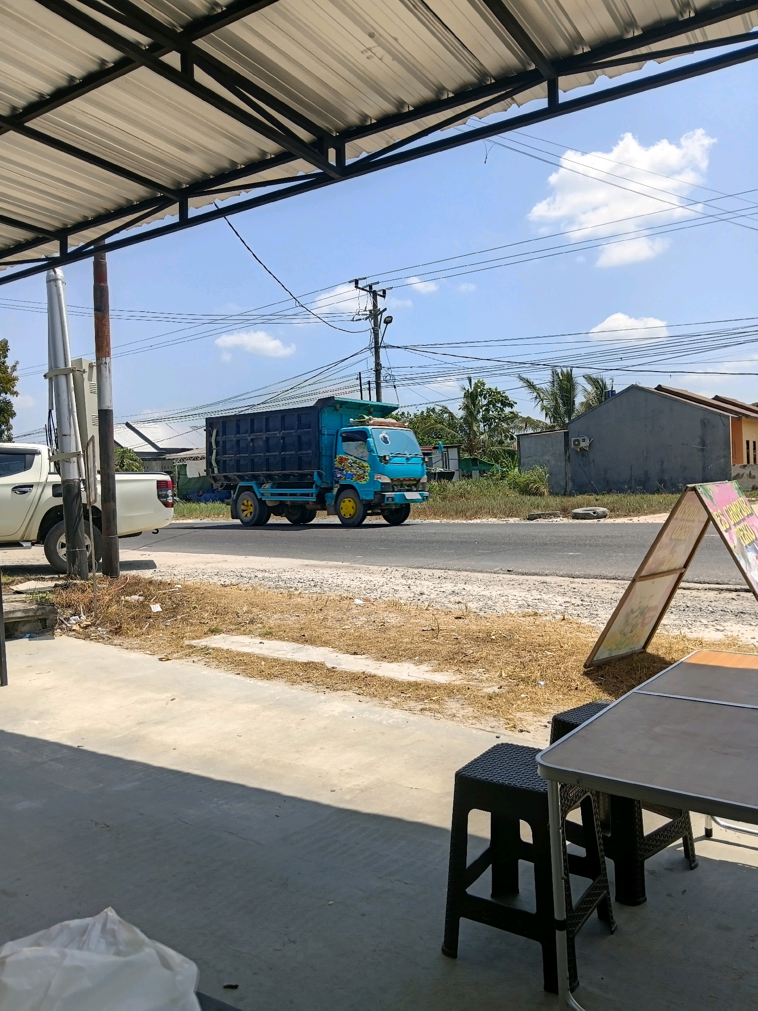 The image captures a street scene in Batu Belaman, Indonesia. In the mid-ground, a bright teal dump truck with yellow wheels is traveling on a paved road. The truck has colorful decals on its side. To the left of the dump truck, a white pickup truck is parked. The road is bordered by dry, sparse vegetation. In the background, there are houses and trees, with utility poles and power lines crisscrossing the sky. A white overhang with a metal frame is visible in the foreground, casting a shadow on the concrete ground. A table and two black stools are set up on the concrete, with an A-frame sign to the right of the table. The sky is clear blue with scattered white clouds. The overall impression is a sunny day in a semi-urban or rural setting.