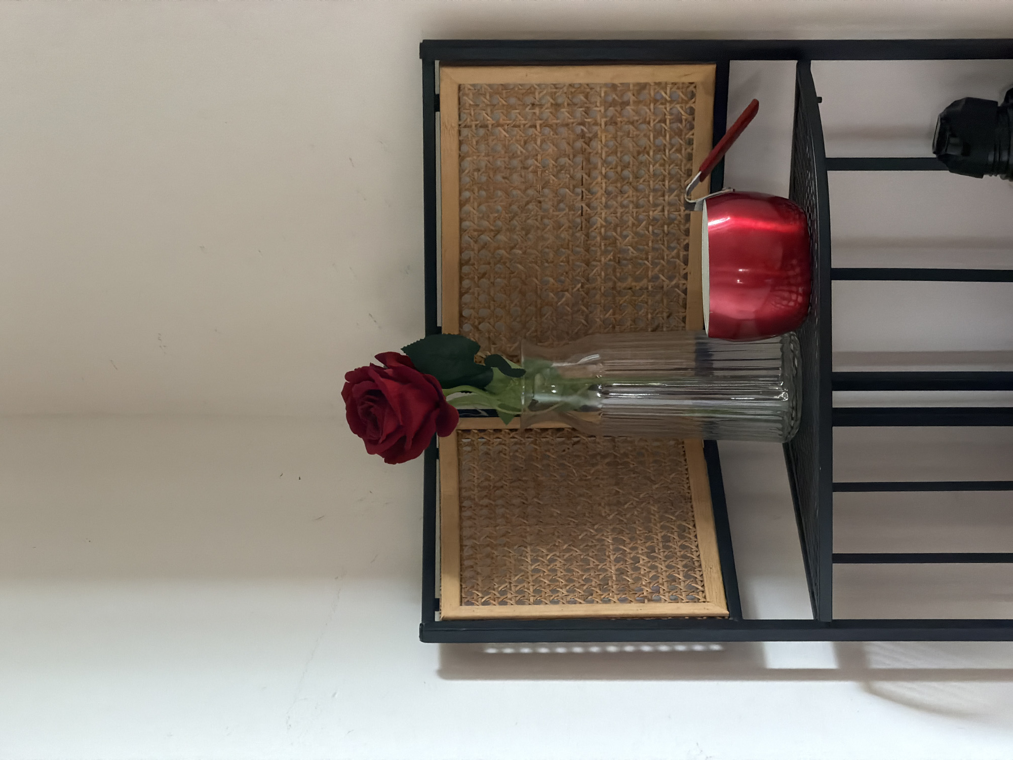 A close-up shot features a metal-framed shelf against a white wall. The shelf has two tiers with woven rattan panels. A long, clear glass vase holding a single red rose rests on the lower tier. A red pot with a long handle is also on the shelf. The lighting is soft and diffused, casting subtle shadows on the wall and the objects. The location is potentially Milan, Italy.
