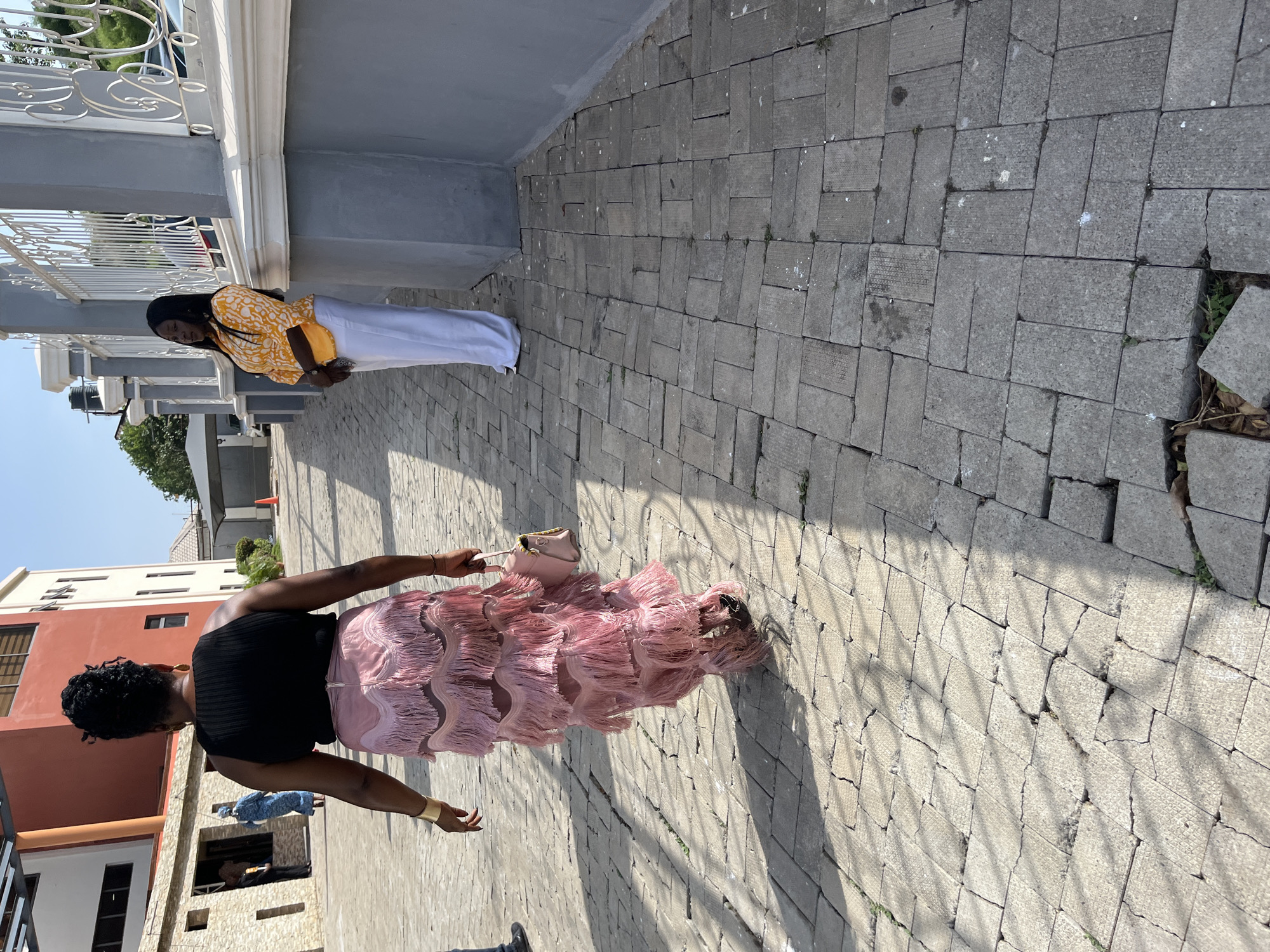 The image shows two women on a paved street in Abuja, Nigeria. One woman is wearing a yellow patterned top and white pants. She is standing near a gray and white building. The second woman is wearing a black top and a pink, fringed skirt. She is holding a pink purse and is walking toward the viewer. The street is made of gray paving stones. Buildings and trees are visible in the background. The sky is clear and blue.