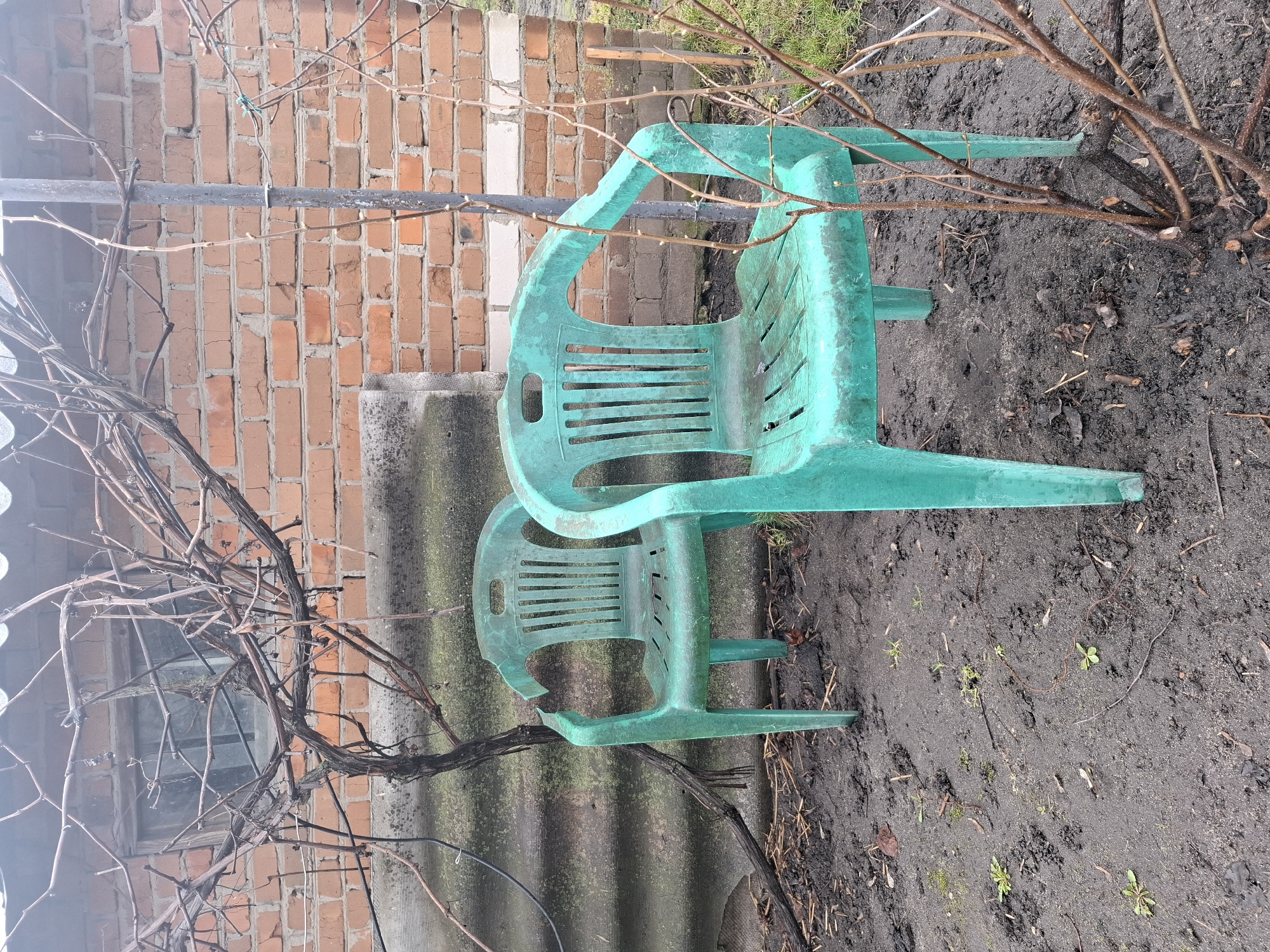 Two green plastic chairs are stacked in a backyard in Khlevnoye, Russia. The background includes a brick wall, a corrugated fence, and bare branches. The ground is muddy.