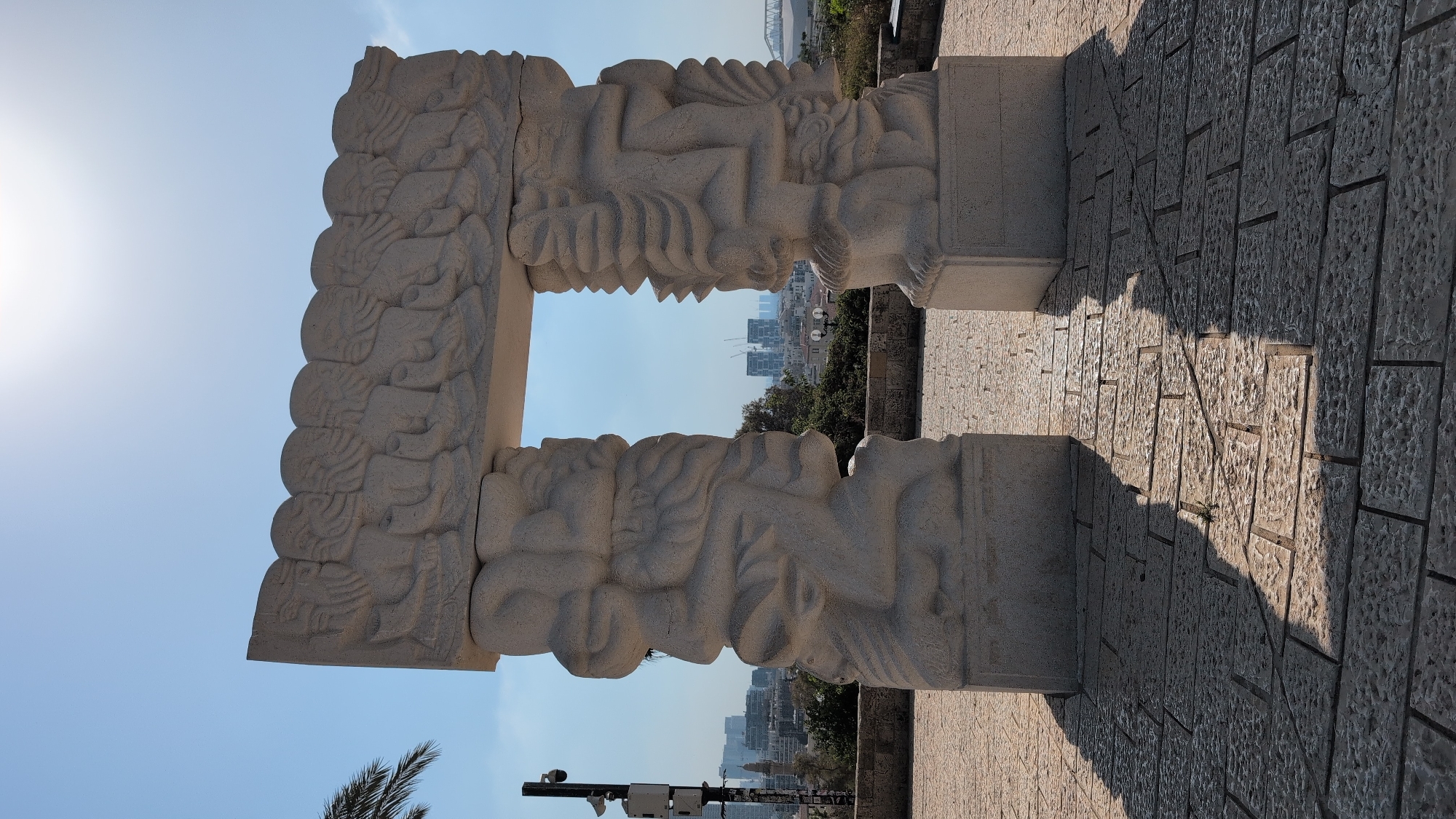 This is a medium shot of a large stone archway sculpture with a cityscape in the background. The sculpture is made of light-colored stone and features intricate carvings on its columns and lintel. The lintel depicts a row of figures with their arms raised. The columns are carved with abstract, swirling forms that suggest human figures or perhaps flowing drapery. The sculpture is set on a cobblestone plaza, and its shadow is cast across the ground.
In the background, beyond the sculpture, is a city skyline with modern buildings and a bridge. The sky is clear and bright blue, with the sun creating a lens flare at the top of the image. The sculpture is situated in a park or elevated viewpoint, as evidenced by the trees and distant buildings. The presence of a security camera on a pole to the left of the sculpture suggests a public area. This sculpture is located at the **Old Jaffa Hilltop**, in **Tel-Aviv, Israel**.
