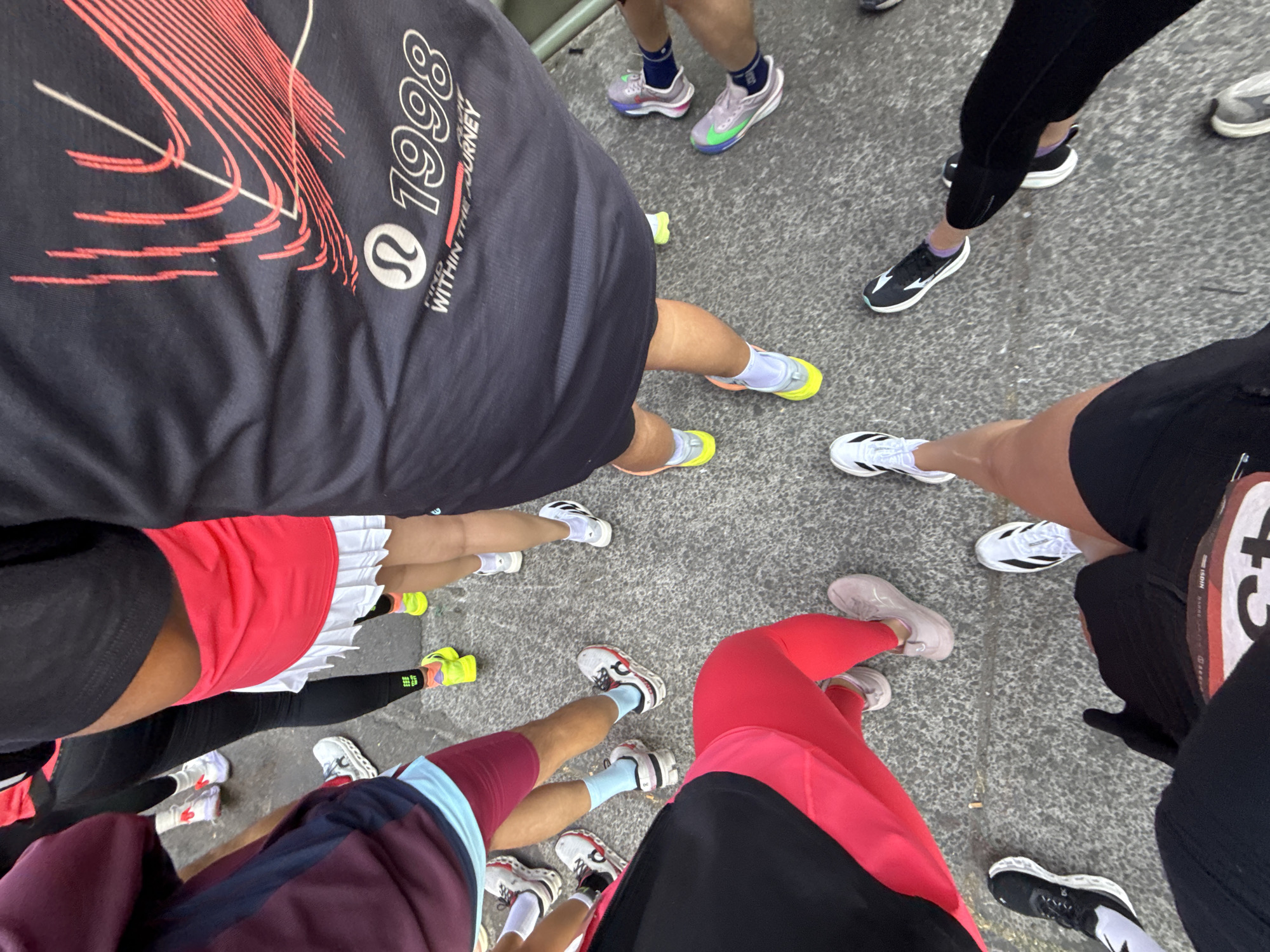 A top-down, wide-angle shot reveals a group of people standing on a gray surface, likely concrete, in what seems to be an outdoor urban setting.
People are visible primarily from the torso down. The scene highlights their diverse athletic wear and running shoes, with a variety of colors and styles. One person is wearing a black shirt with graphics and the number "1998." Another person is wearing a red and white tennis skirt, and black leggings. One person is wearing a pink top with black shorts.
The focus is primarily on the lower bodies of the group, emphasizing the variety of athletic footwear and attire. The individuals are grouped closely together, suggesting they are part of an event or gathering.
Given the "within the journey" and the Lululemon logo on the shirt, it is most likely they are waiting for a running event to start.
It is not possible to determine if this is in Mexico City.