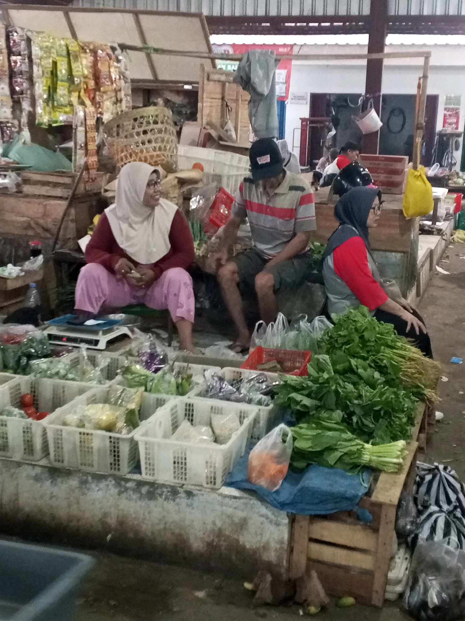 The image depicts an outdoor market scene in Gentan, Indonesia. Three individuals are visible. On the left, a woman wearing a white hijab and a maroon top is sitting and looking down, seemingly engaged with something in her hands. She is positioned in front of a weighing scale and surrounded by various vegetables displayed in white plastic crates. In the center, a man wearing a black cap and a striped polo shirt is also seated, facing towards the left. To his right, a woman wearing a dark hijab and a red and gray long-sleeved shirt is seated, looking to her right. The market stall features a variety of fresh produce, including leafy greens and what appear to be tomatoes and carrots, along with plastic bags and baskets. The overall environment suggests a bustling local marketplace.