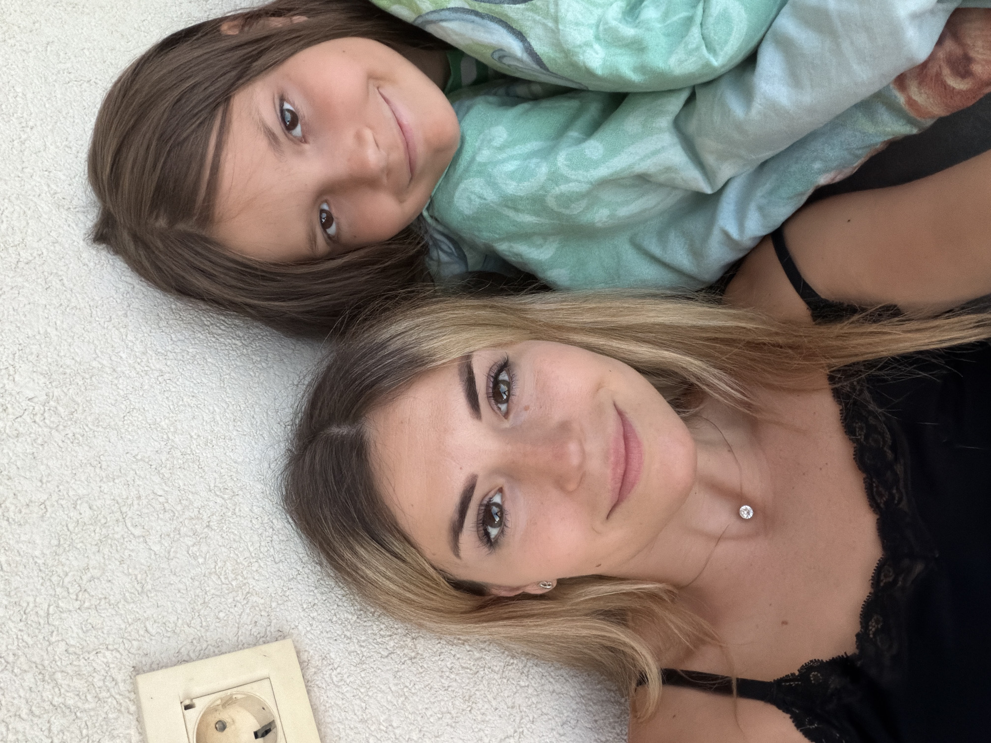 A close-up, overhead shot shows two females, one adult and one child, posing for a selfie against a textured, off-white wall. To the left, an electrical outlet is visible. The adult, on the left, has blonde hair and wears a black lace-trimmed top and a delicate necklace with a single diamond. The child, on the right, has long brown hair and is wrapped in a green blanket with a pattern of white and light blue, featuring some faint orange shapes. Both are looking towards the camera with subtle smiles. The background suggests an outdoor setting, potentially a terrace or balcony, as the wall appears to be exterior. The overall scene evokes a relaxed and casual atmosphere. The image was taken in Budva, Montenegro.