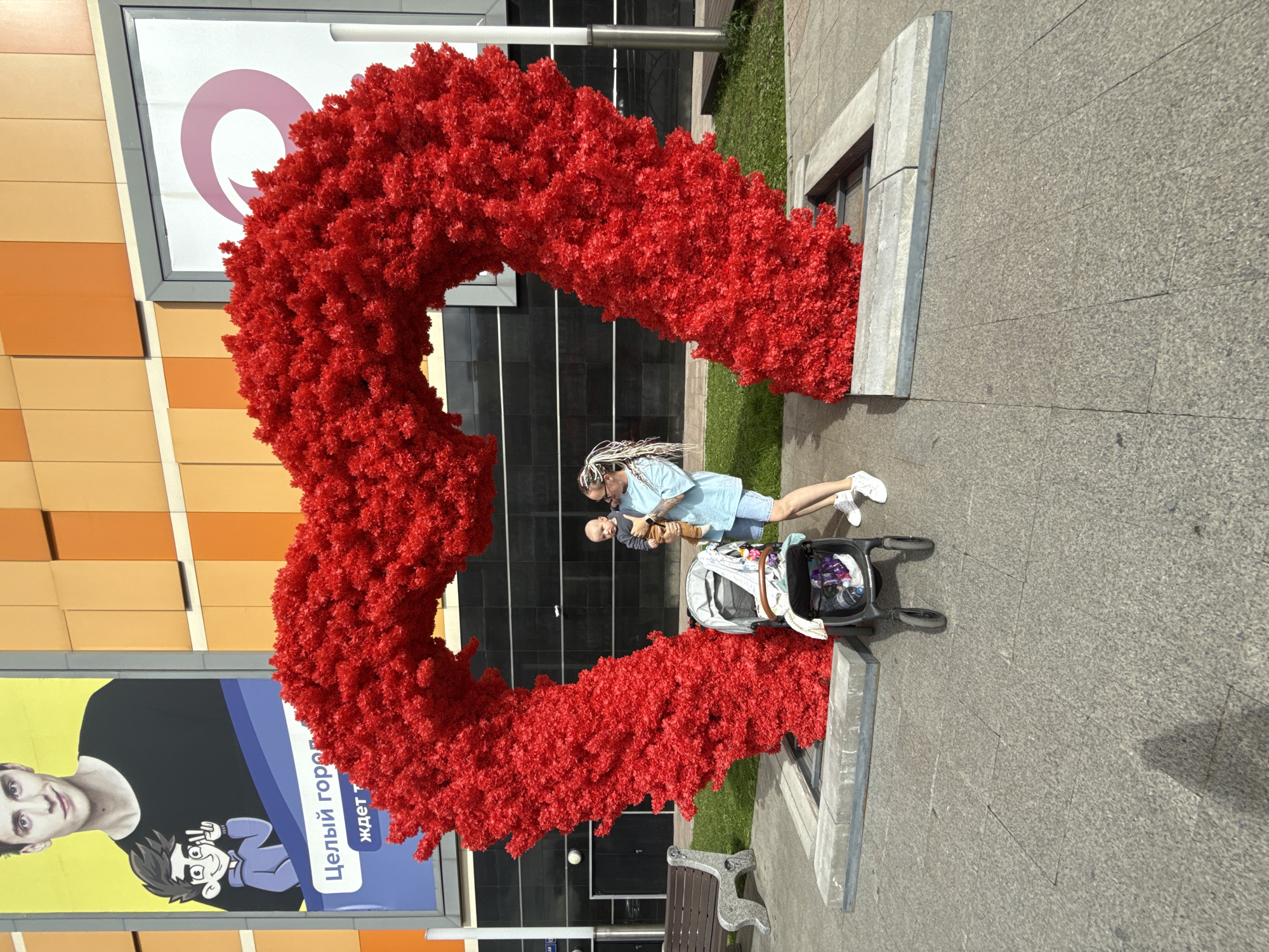 A woman with blonde braided hair stands next to a red heart-shaped archway in what appears to be a public outdoor space. She is holding a baby. A stroller is parked next to her. In the background, there are billboards and a building with an orange facade. One billboard features a young man and text in Russian that reads "Целый город ждет тебя" (The whole city awaits you). Another billboard displays a large pink circular logo. The ground is paved with stone tiles. The scene suggests a location within Moscow, Russia, possibly a shopping center or entertainment district, given the presence of billboards and modern architecture.