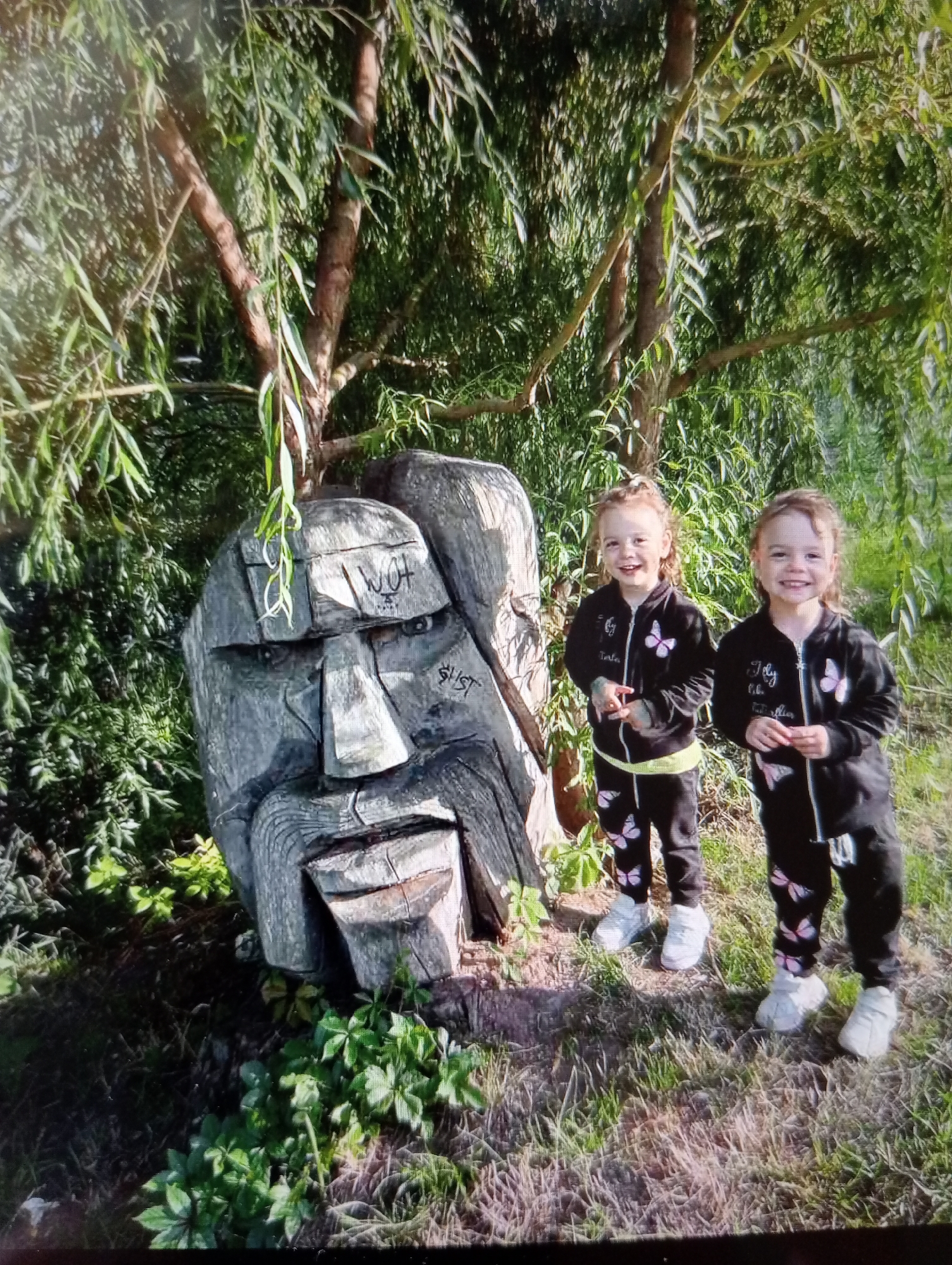An outdoor, eye-level, full shot shows two young Caucasian girls standing beside a large wooden sculpture of a face. The sculpture is gray and appears weathered. It has rough carvings for eyes, a nose, and a mouth. There are some markings on the sculpture's forehead that appear to be graffiti. The sculpture is positioned under the branches of a large tree with long, drooping leaves, possibly a willow. The ground is covered with grass and some leafy plants. The two girls are dressed in matching black tracksuits with pink butterfly patterns and white sneakers. They are both smiling at the camera. The girl on the left has her hands near her chest, while the girl on the right is holding a small red object in her right hand. The background is filled with greenery from the tree and other vegetation. The lighting suggests it is daytime, with sunlight filtering through the leaves.