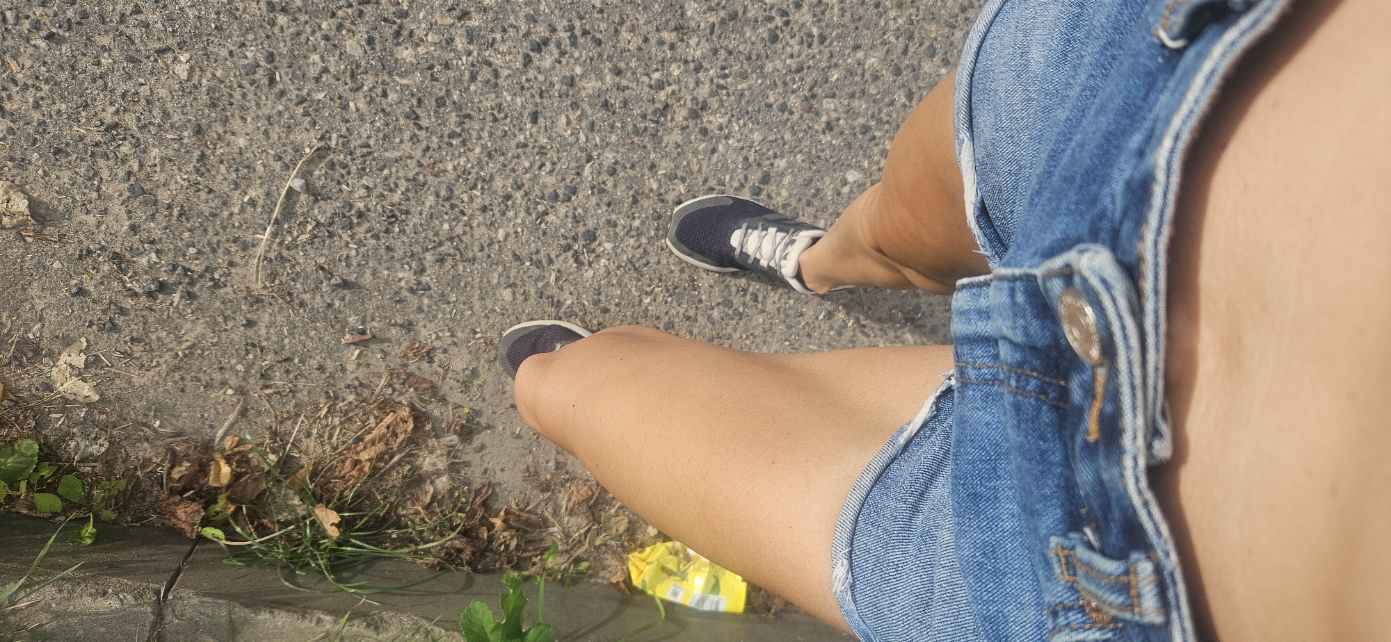 The image is an overhead shot of a person's legs and feet wearing denim shorts and athletic shoes, standing on a textured asphalt surface. There is dried foliage and some green vegetation along the right side of the frame, adjacent to a concrete curb. A bright yellow wrapper is visible on the ground near the curb. The lighting suggests it is daytime. There is no discernible information in the image that would allow for the identification of the location as Vitsebsk, Belarus.