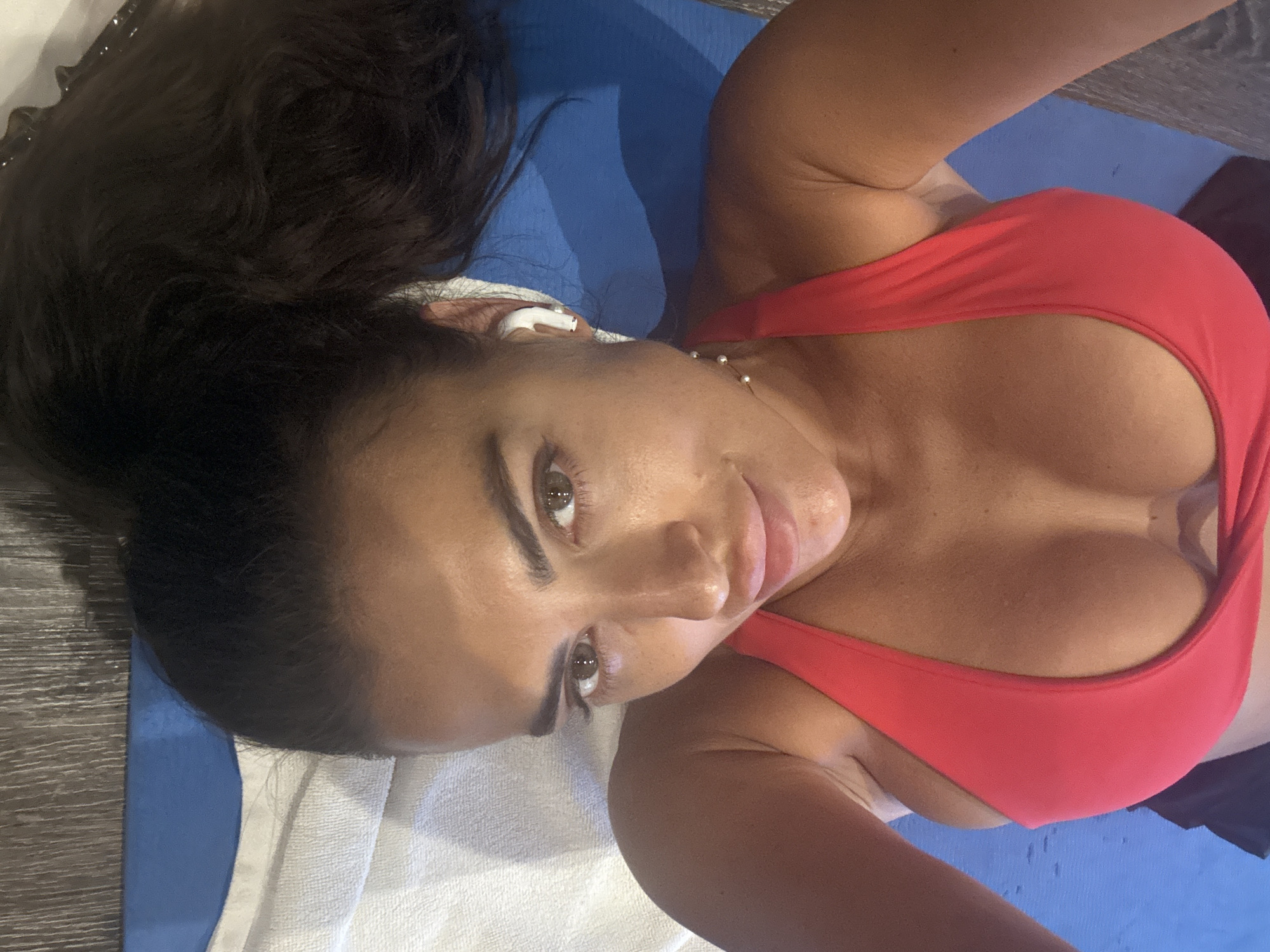 A woman lies on a blue yoga mat and a white towel. She is wearing a red sports bra. She has dark hair pulled up and an earbud in her left ear. She is tan. There are light-colored beads around her neck. It is indoors. Location of North Kuta, Indonesia is not confirmed by the visual data in the image.