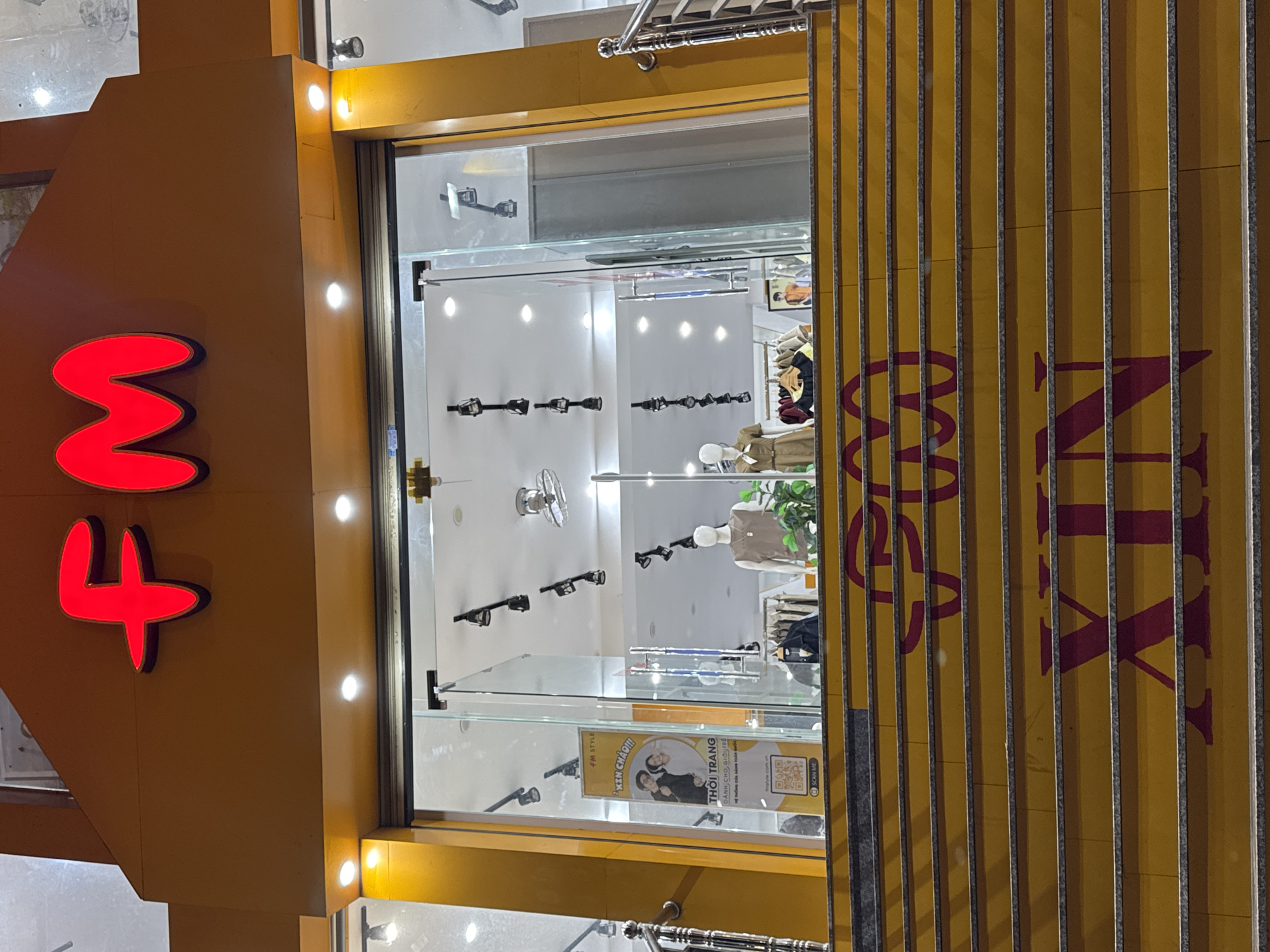 The image captures a storefront in Da Lat, Vietnam, during what appears to be nighttime or late evening due to the artificial lighting. The storefront features a prominent yellow facade with vertical metal grating on the right side, behind which large, dark red lettering spells out "FM SA". On the left side of the facade, large, illuminated red shapes resembling the numbers "3" and "4" are visible.
The main focus is the glass-enclosed storefront, offering a view into the interior. Inside, mannequins are displayed, dressed in clothing, suggesting it is a clothing store. The interior is brightly lit with spotlights, illuminating display shelves and racks holding various items, possibly accessories like sunglasses, which are mounted on the glass partitions. A promotional banner is visible inside, featuring two individuals and text in Vietnamese, including "XIN CHÀO!!!" (Hello!!!) and "THỜI TRANG DÀNH CHO GIỜI TRẺ" (Fashion for Young People), along with a QR code labeled "SCAN ME". The overall impression is that of a modern retail establishment, likely a fashion boutique catering to a younger demographic, situated in a commercial area of Da Lat. The lighting creates a somewhat dynamic and inviting atmosphere.