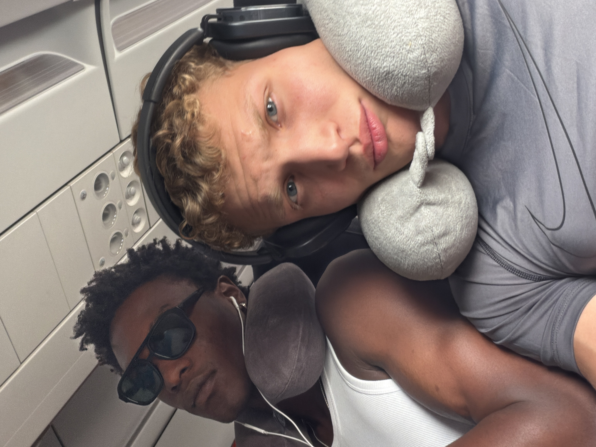 Two young men are seated next to each other in what appears to be an airplane cabin, likely on a flight. The man on the left, with dark skin and curly hair, wears sunglasses and a white tank top, with a neck pillow supporting his head. He has earbuds in and is looking upwards. The man on the right, with fair skin and curly blonde hair, wears headphones and a grey t-shirt. He is looking directly at the camera, with a grey neck pillow around his neck. The overhead cabin lights and interior of the plane are visible in the background. The image does not provide any specific information or context related to Marrakesh, Morocco.